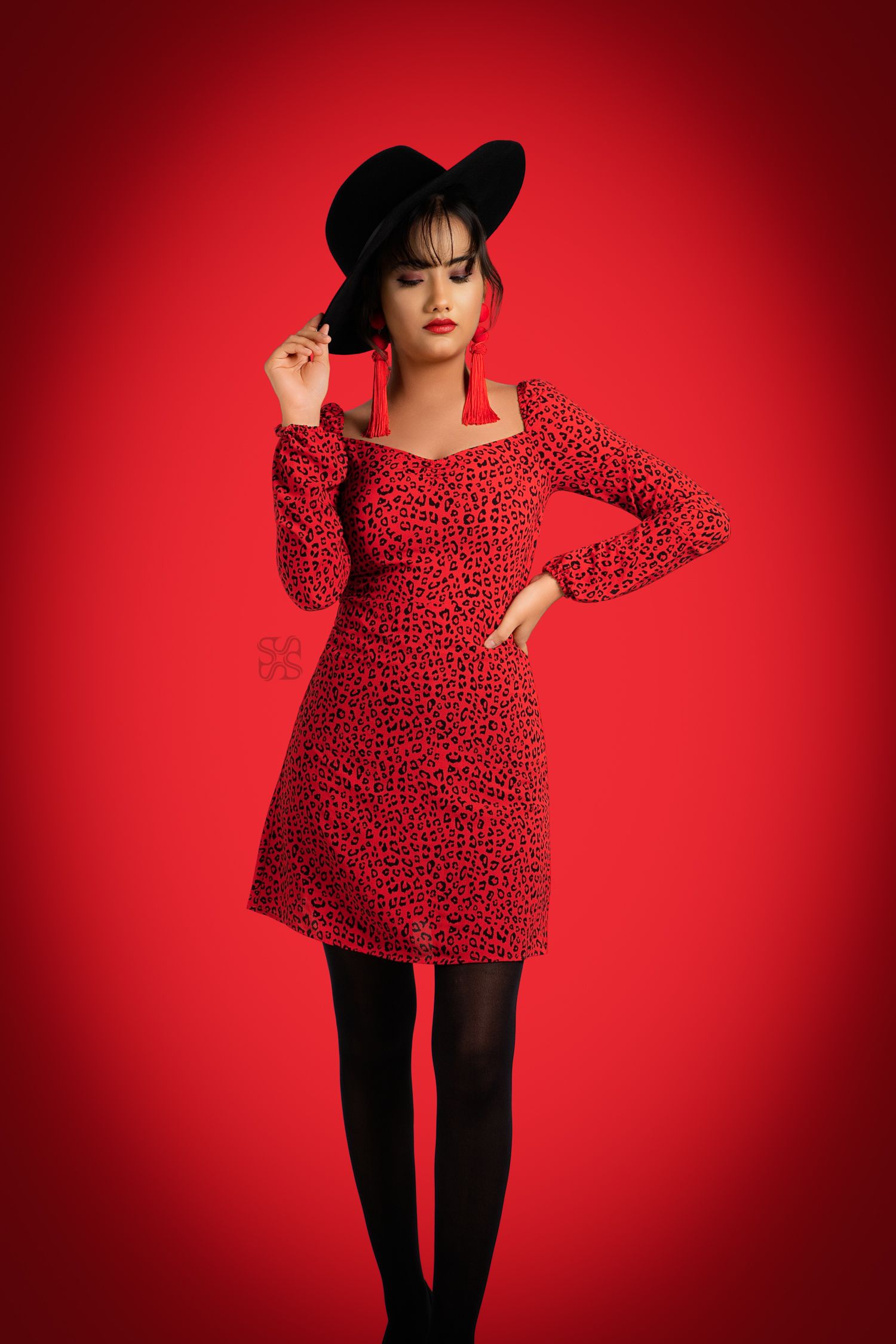 Woman in red leopard print dress and black hat posing on red background.