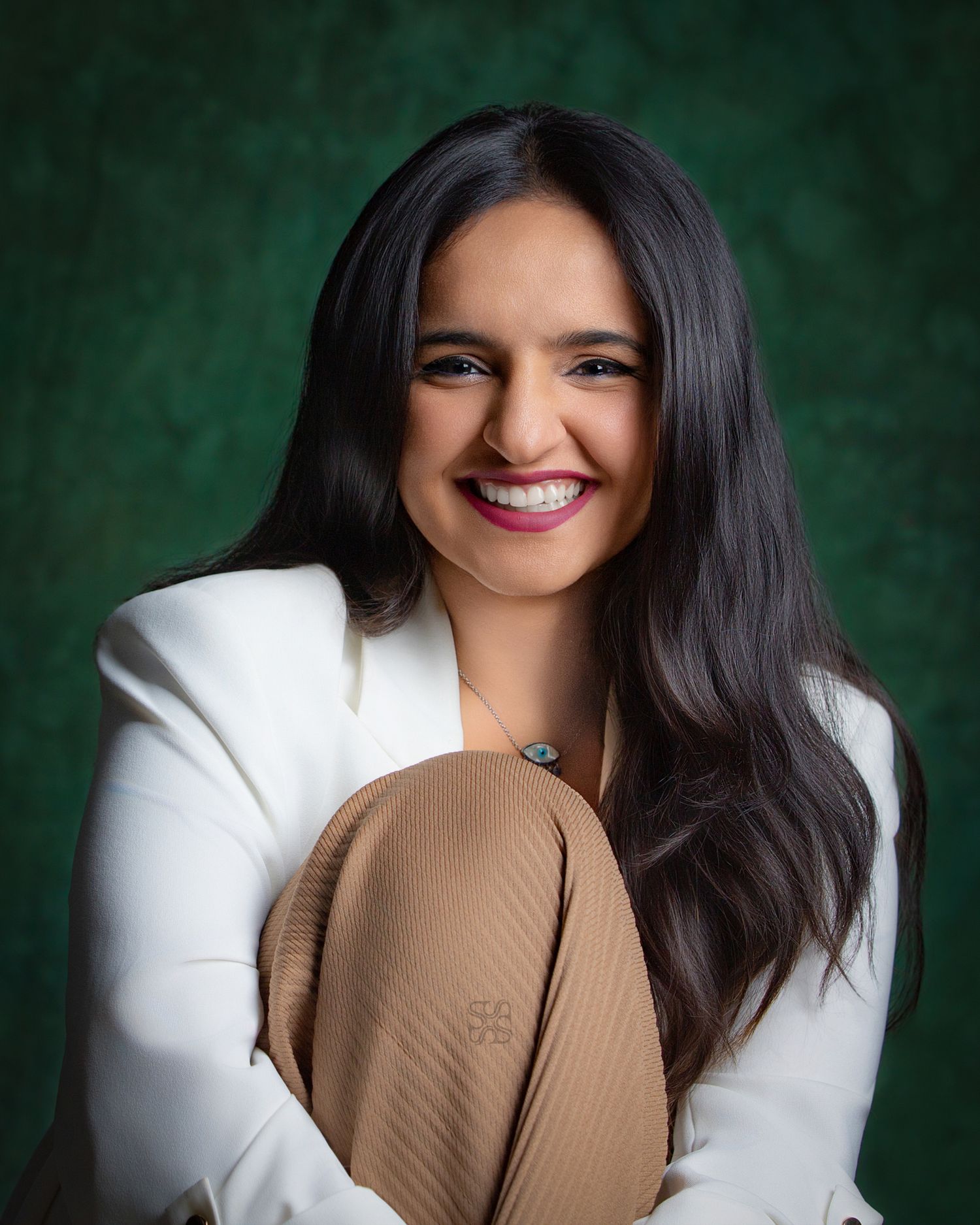 Smiling young woman with long dark hair in white blazer and tan pants, posing against green background.