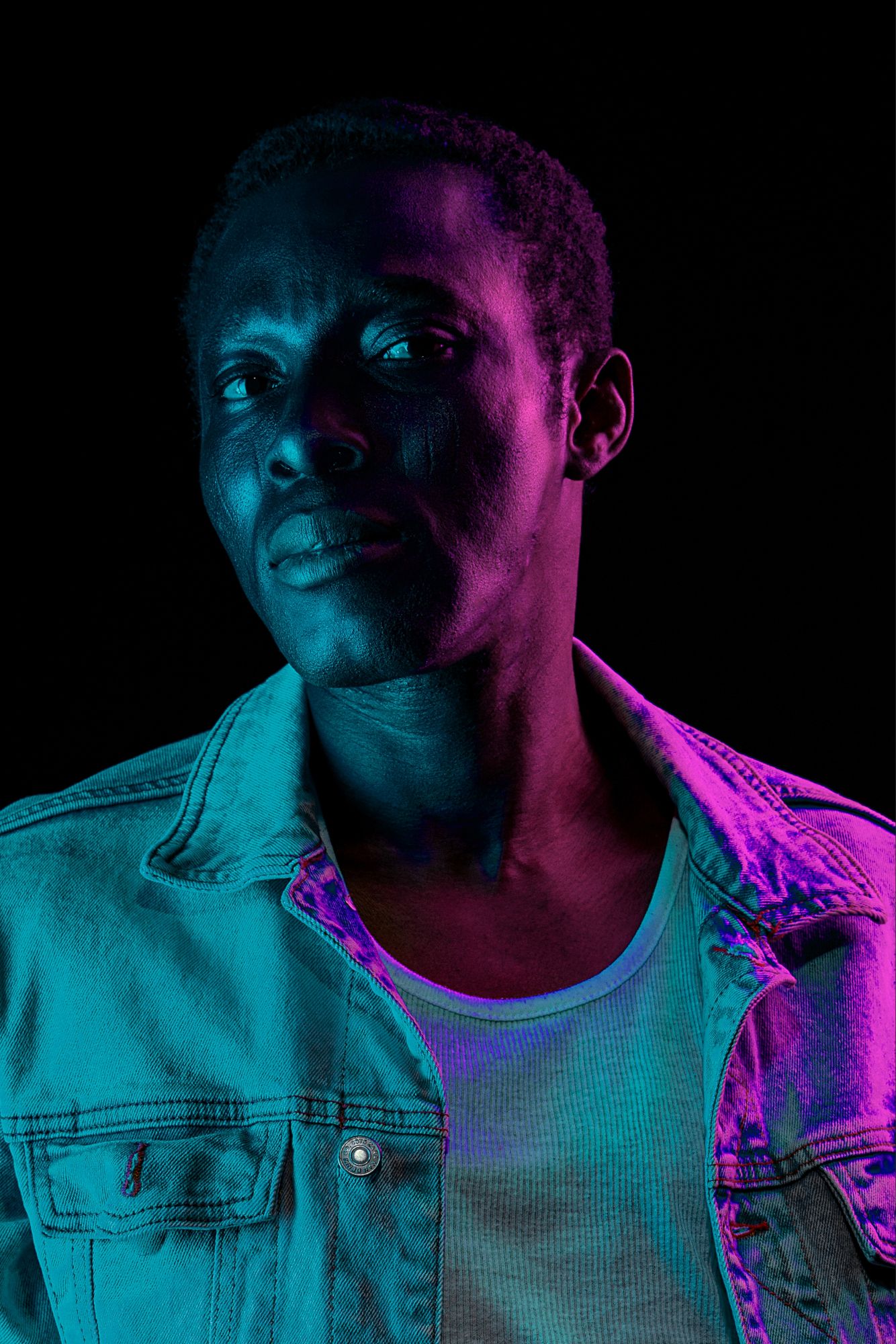 Portrait of a young Black man in a denim jacket under dramatic neon blue and purple studio lighting.