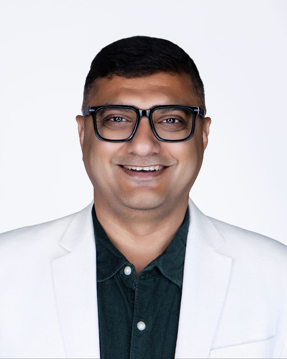 Smiling man wearing black glasses and white blazer over dark green shirt poses for professional headshot.
