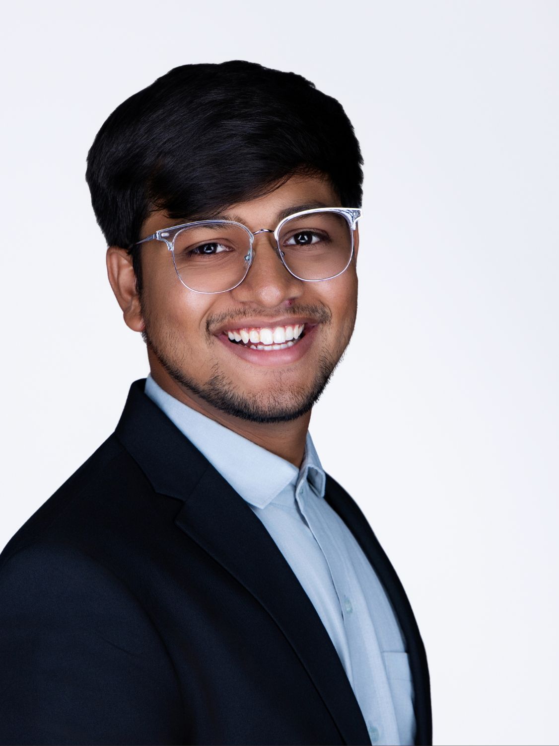 Young man smiling in glasses and dark blazer, professional headshot on white background.
