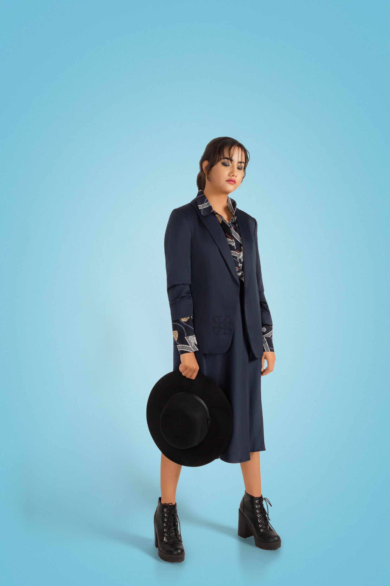 Woman in navy blazer suit and black boots holding a wide-brim hat on blue background.