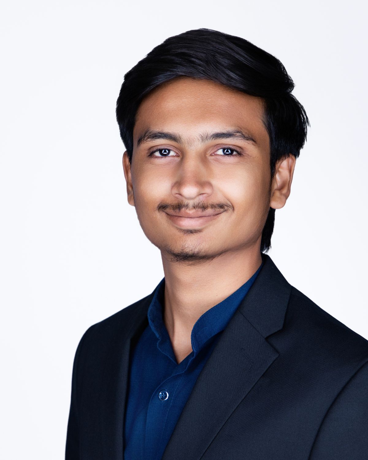 Young professional man in dark blazer and blue shirt smiling confidently against white background.