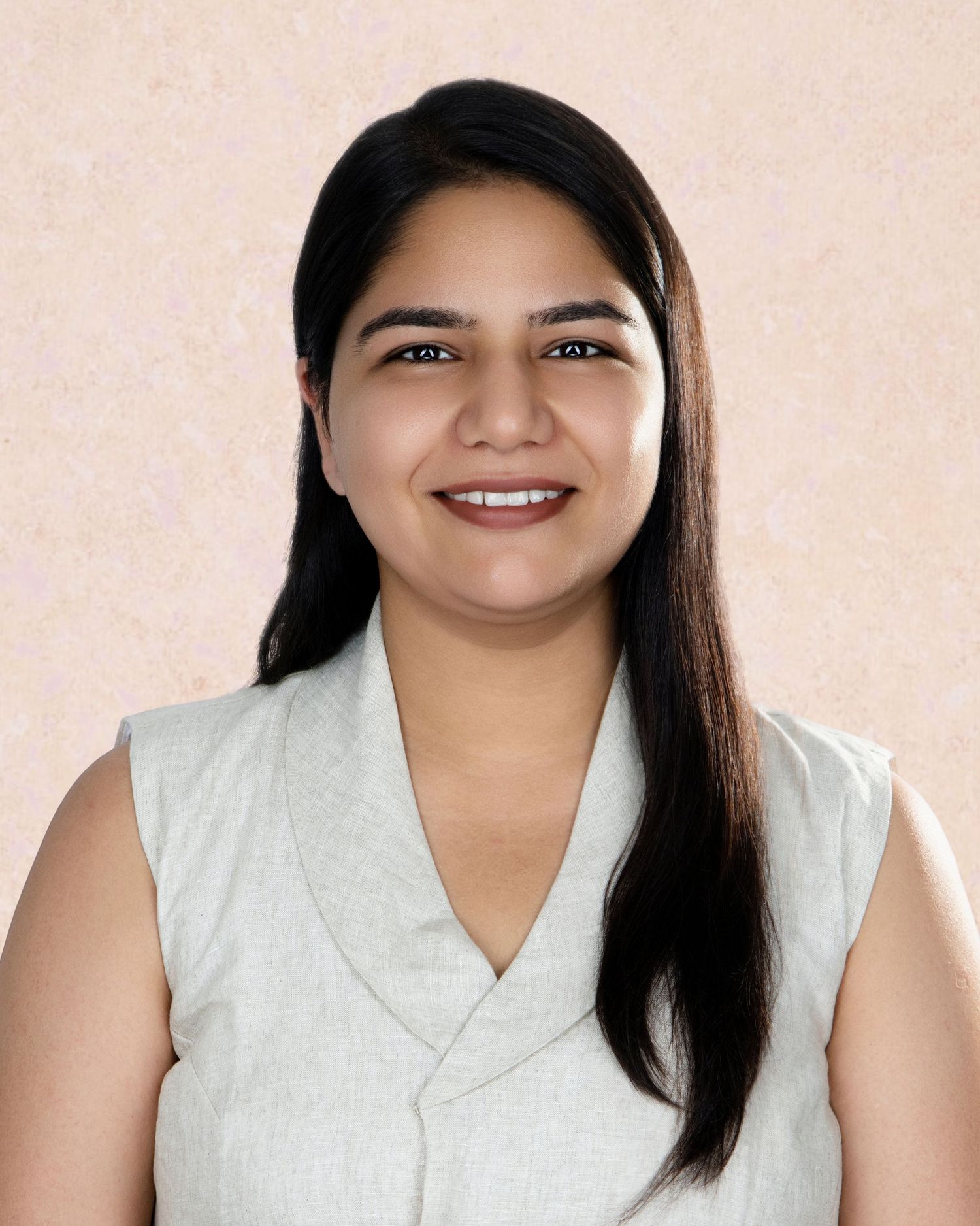 Smiling young woman with long dark hair wearing a sleeveless gray top against a beige background.