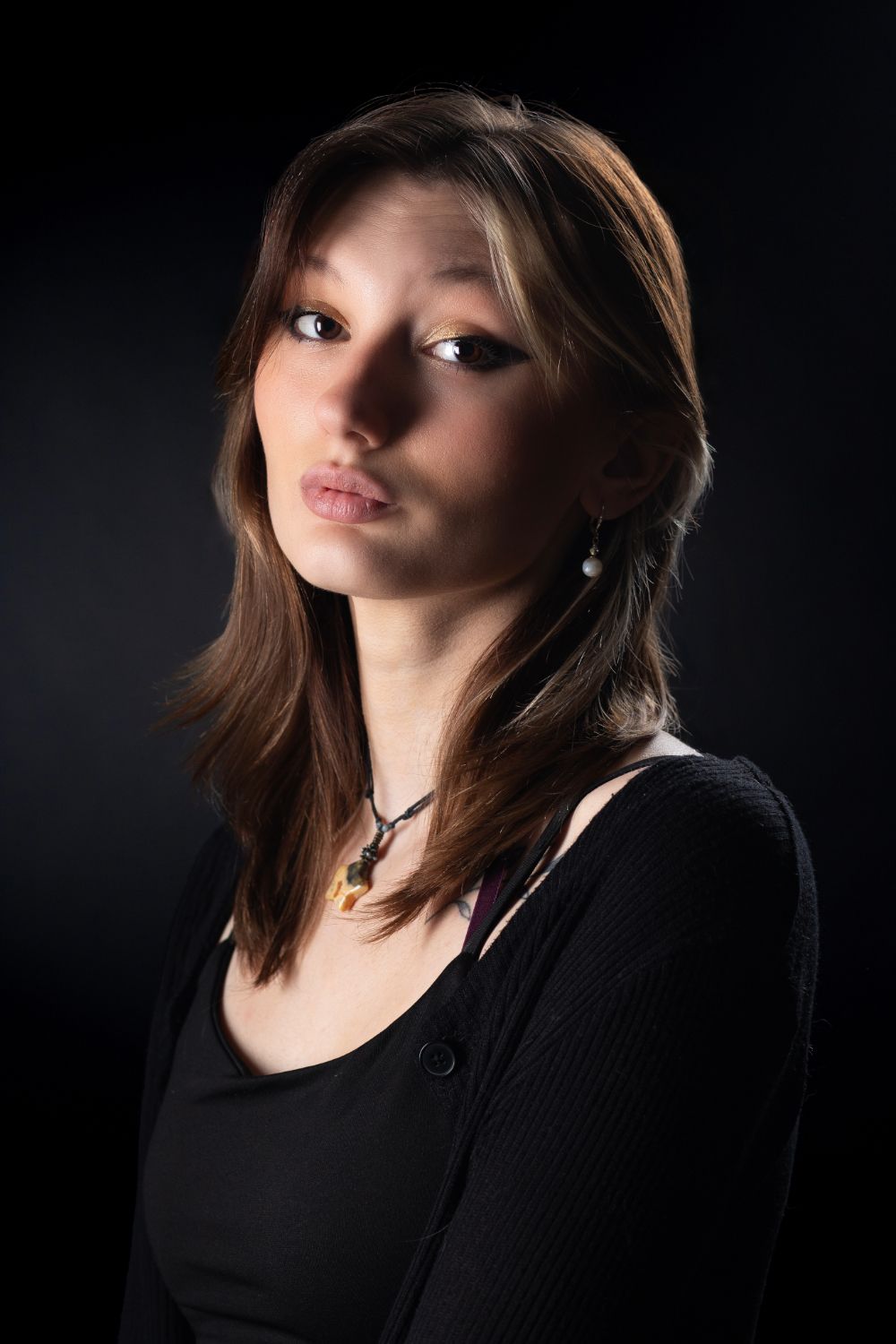 Young woman with brown hair wearing black cardigan and necklace poses against dark background.