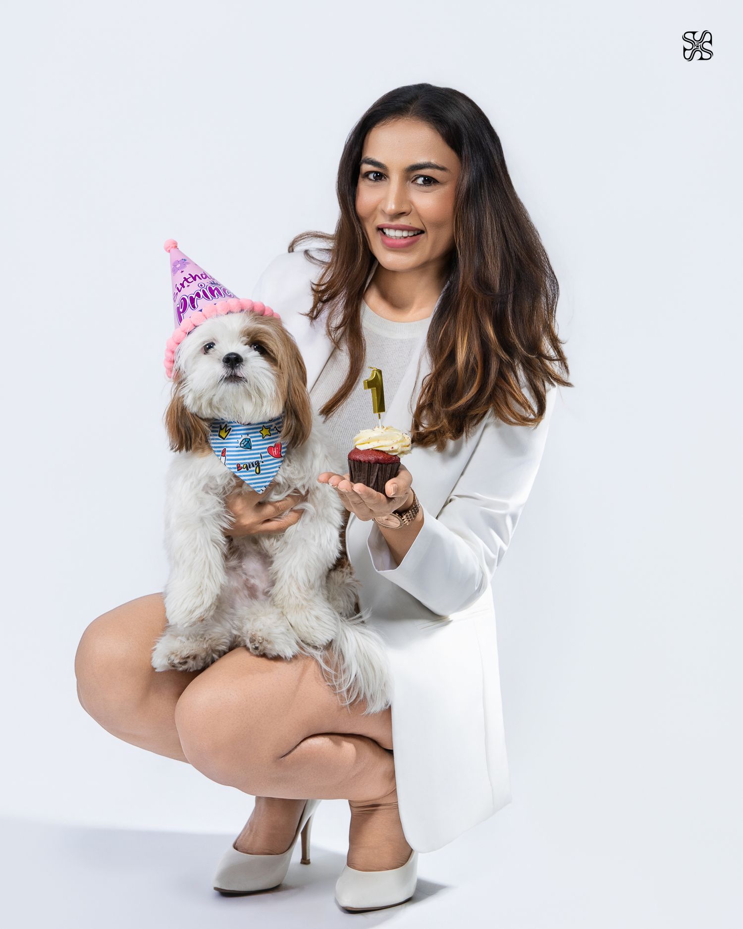 Woman in white outfit crouching with a Shih Tzu dog wearing a birthday hat, holding a cupcake with a number 1 candle.