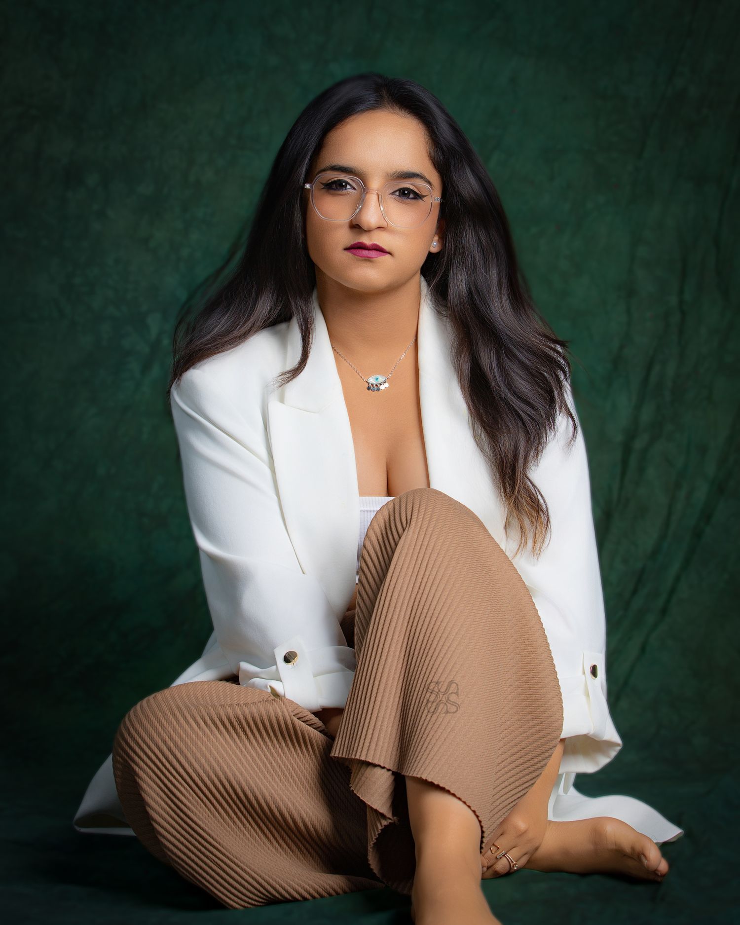 Woman in white blazer and tan pleated pants sits gracefully against a dark green backdrop.