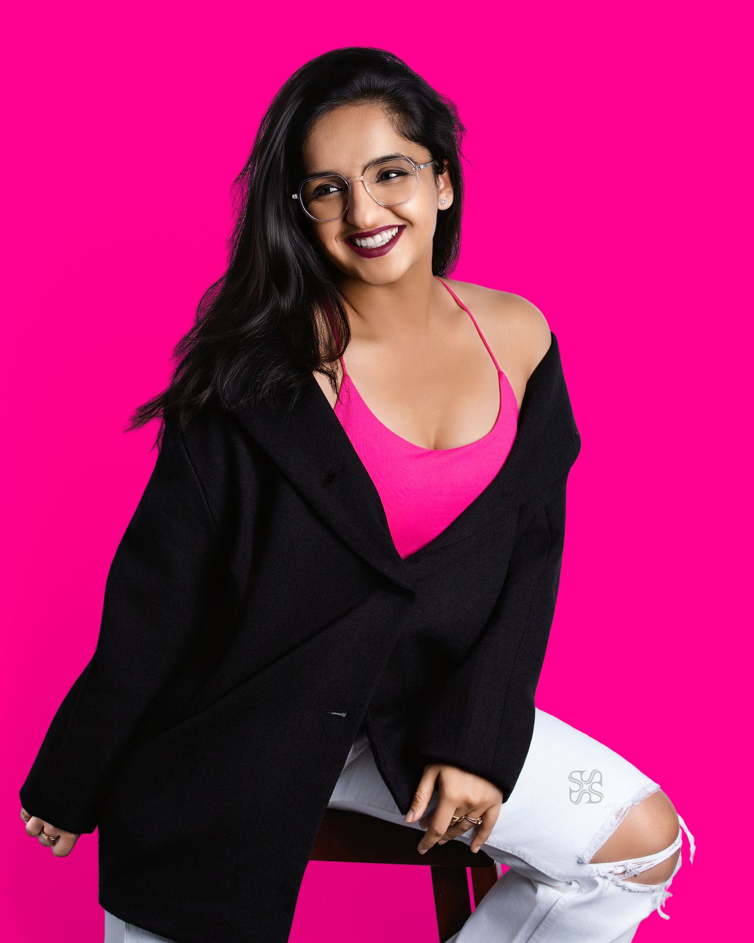 Woman in pink top, black blazer, and white ripped jeans posing confidently against a vibrant pink background.