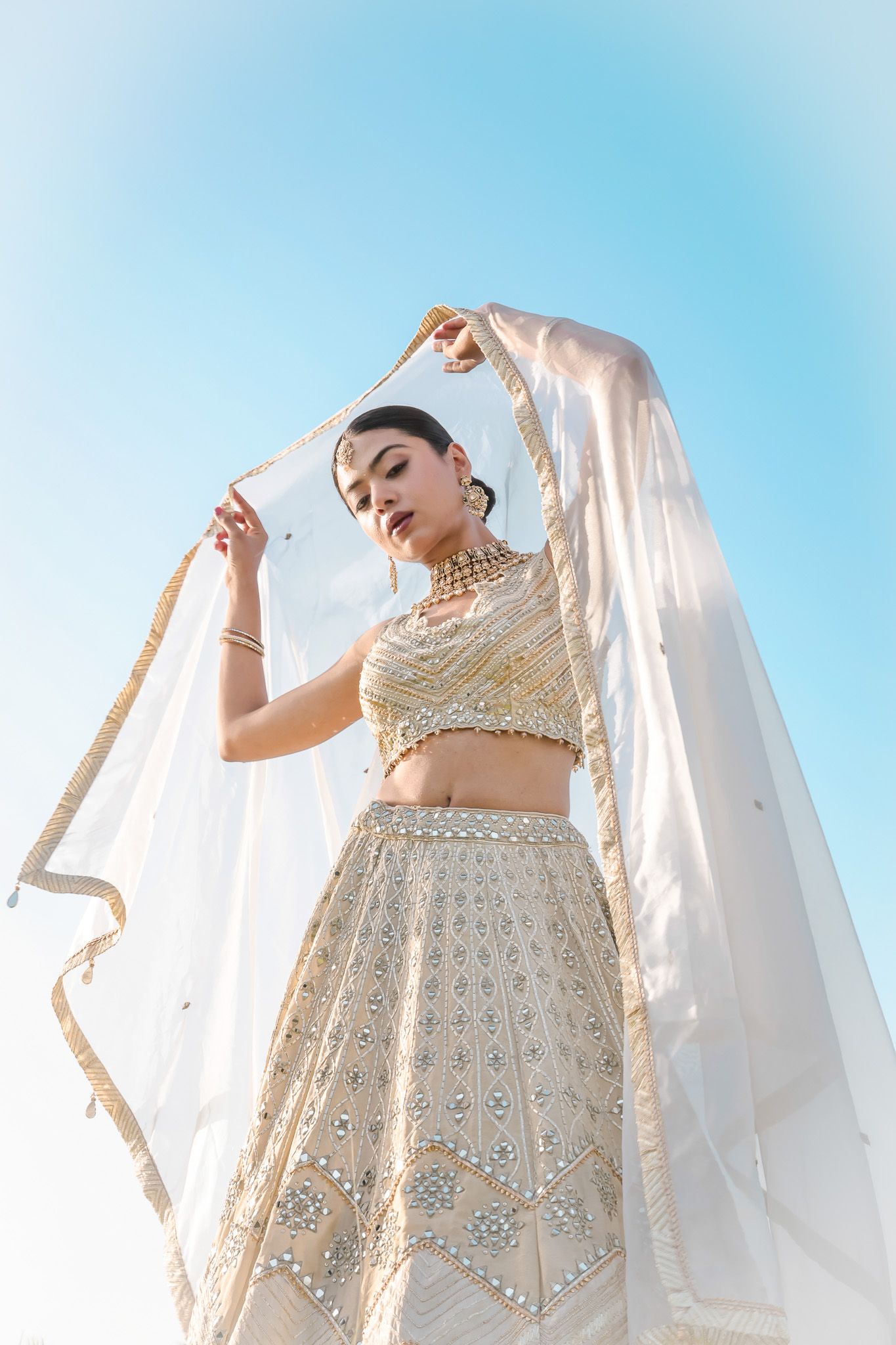 A bride in white and gold embroidered lehenga holds her dupatta against a blue sky.