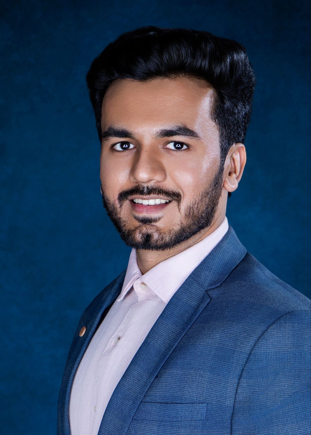 Young professional man in blue suit smiling confidently against dark blue background, formal business portrait.