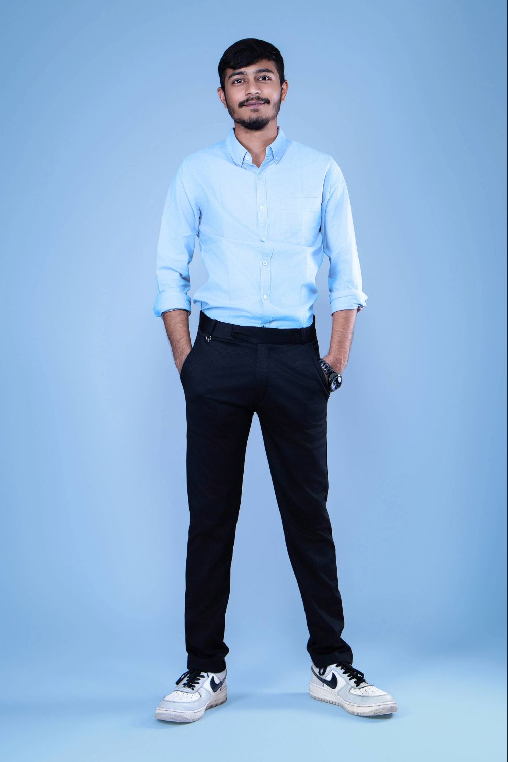 Person in light blue button-down shirt and dark pants poses against blue backdrop with sneakers.
