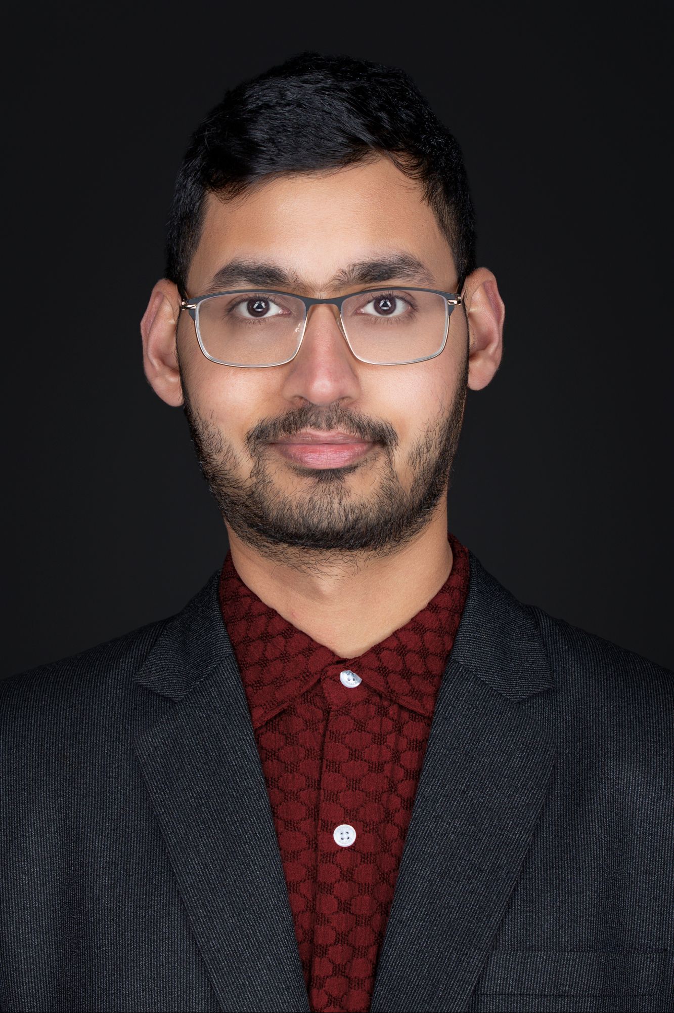 Studio portrait of person in burgundy patterned shirt and dark blazer wearing glasses.
