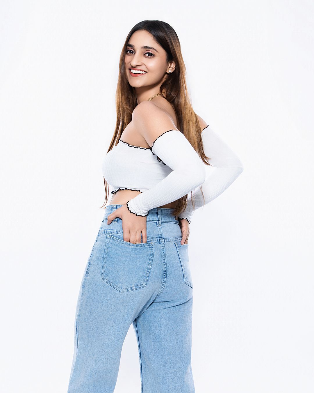 Young woman smiling in white off-shoulder crop top and light blue wide-leg jeans posing against white background.