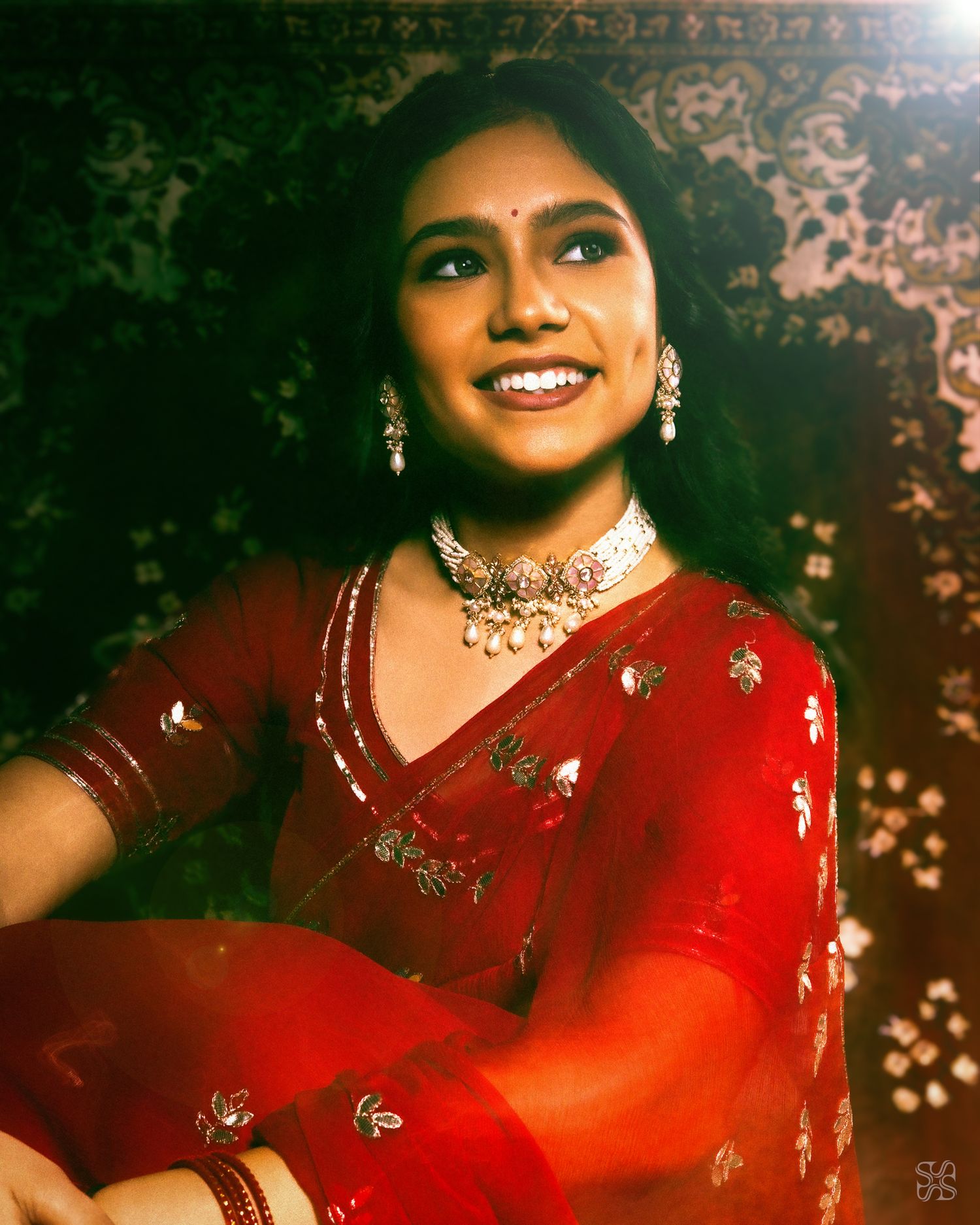 Young woman in red embroidered saree and silver choker necklace smiling gracefully against a dark floral background.