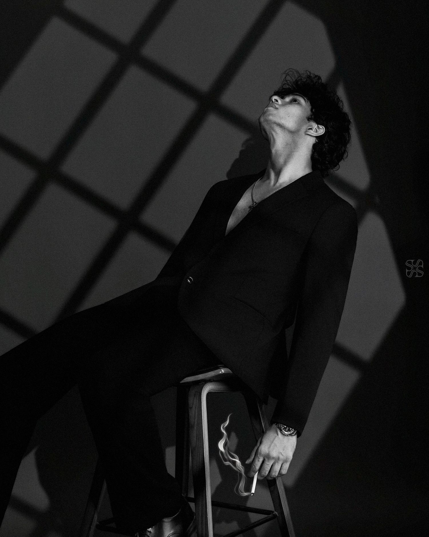 Black and white photo of a young man in a suit sitting on a stool, head tilted back, with dramatic window light shadows.