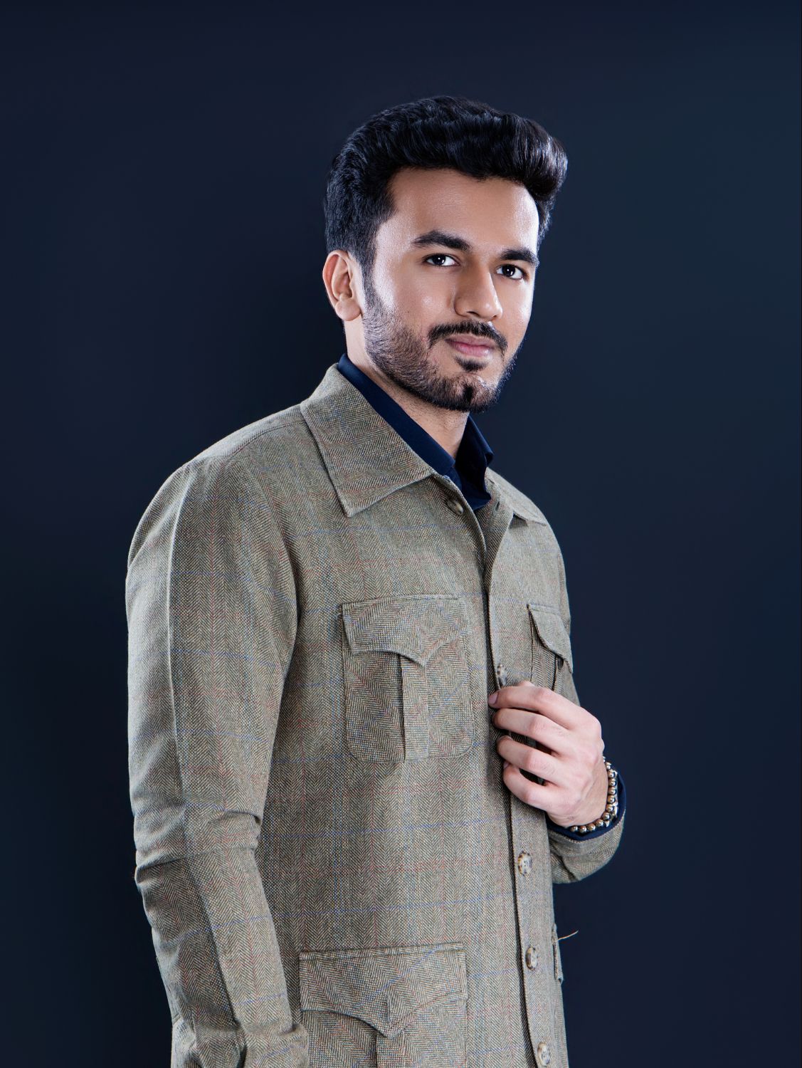 Young man in olive jacket posing confidently against dark background, wearing a beaded bracelet.