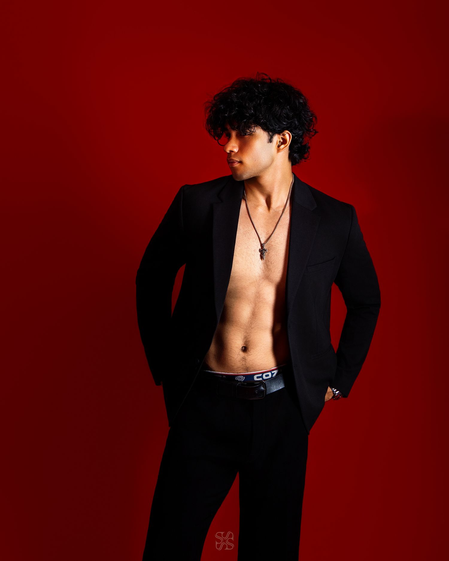 Young man with curly hair in open black blazer posing against bold red background.