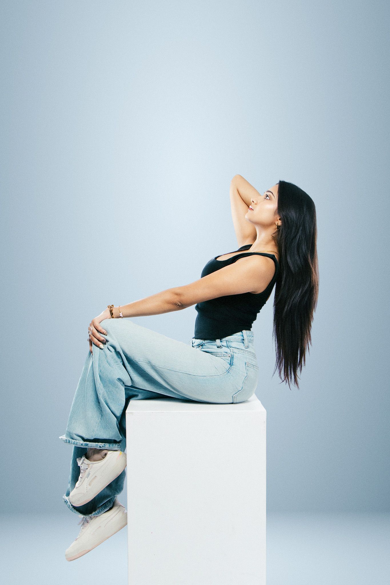 Woman in black top and wide-leg jeans sits on white cube, posing with hand behind head against blue background.