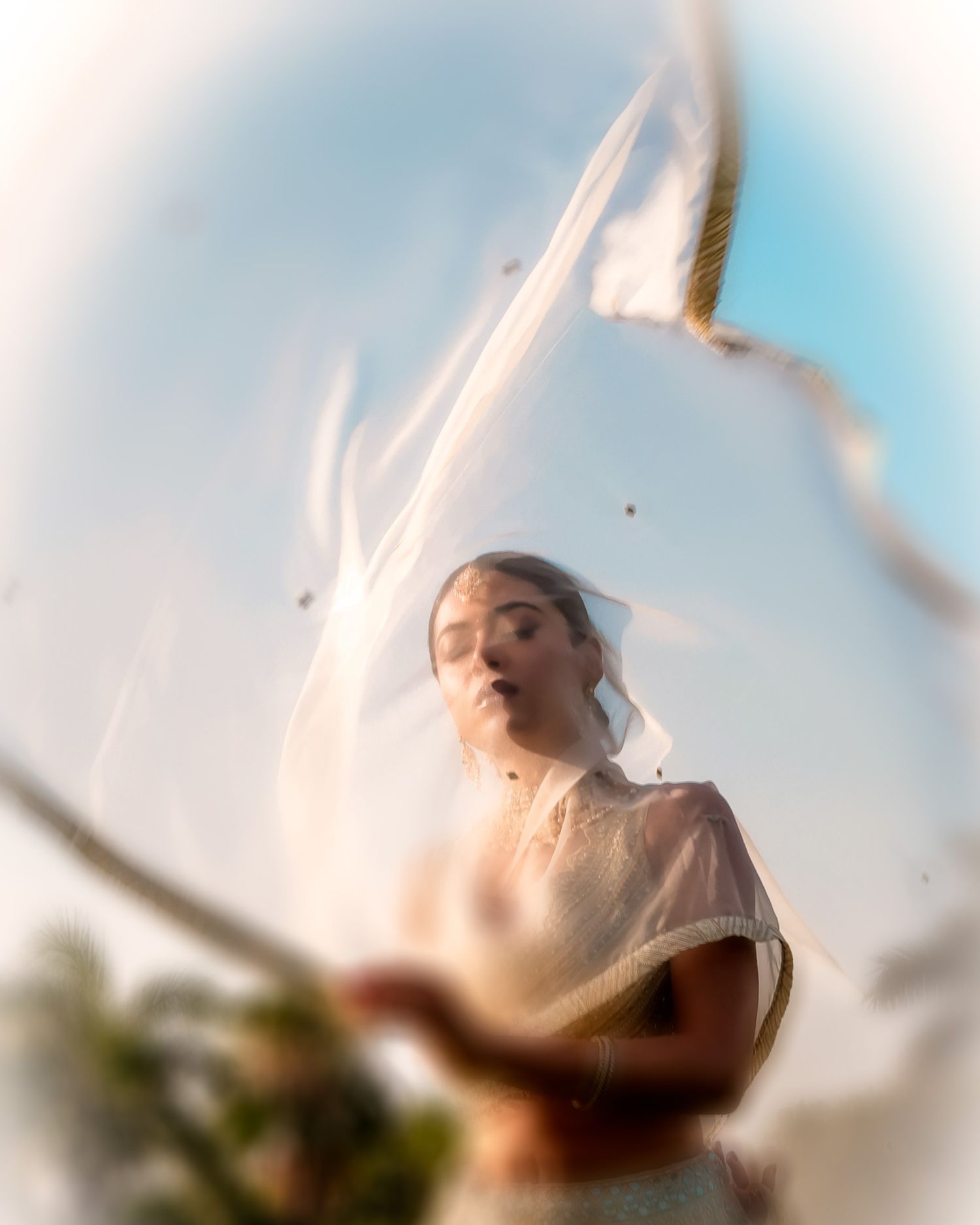 A bride in a white veil and dress stands outdoors, captured through a dreamy fisheye lens with soft bokeh and motion blur.