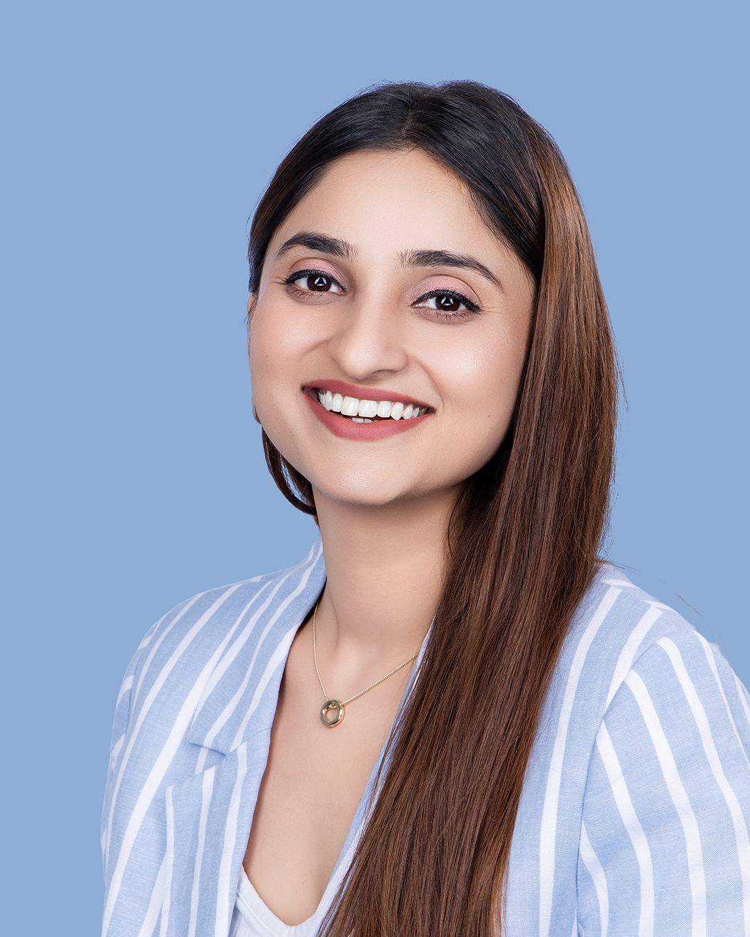 Smiling young woman with long brown hair wearing a striped blazer poses against a light blue background.