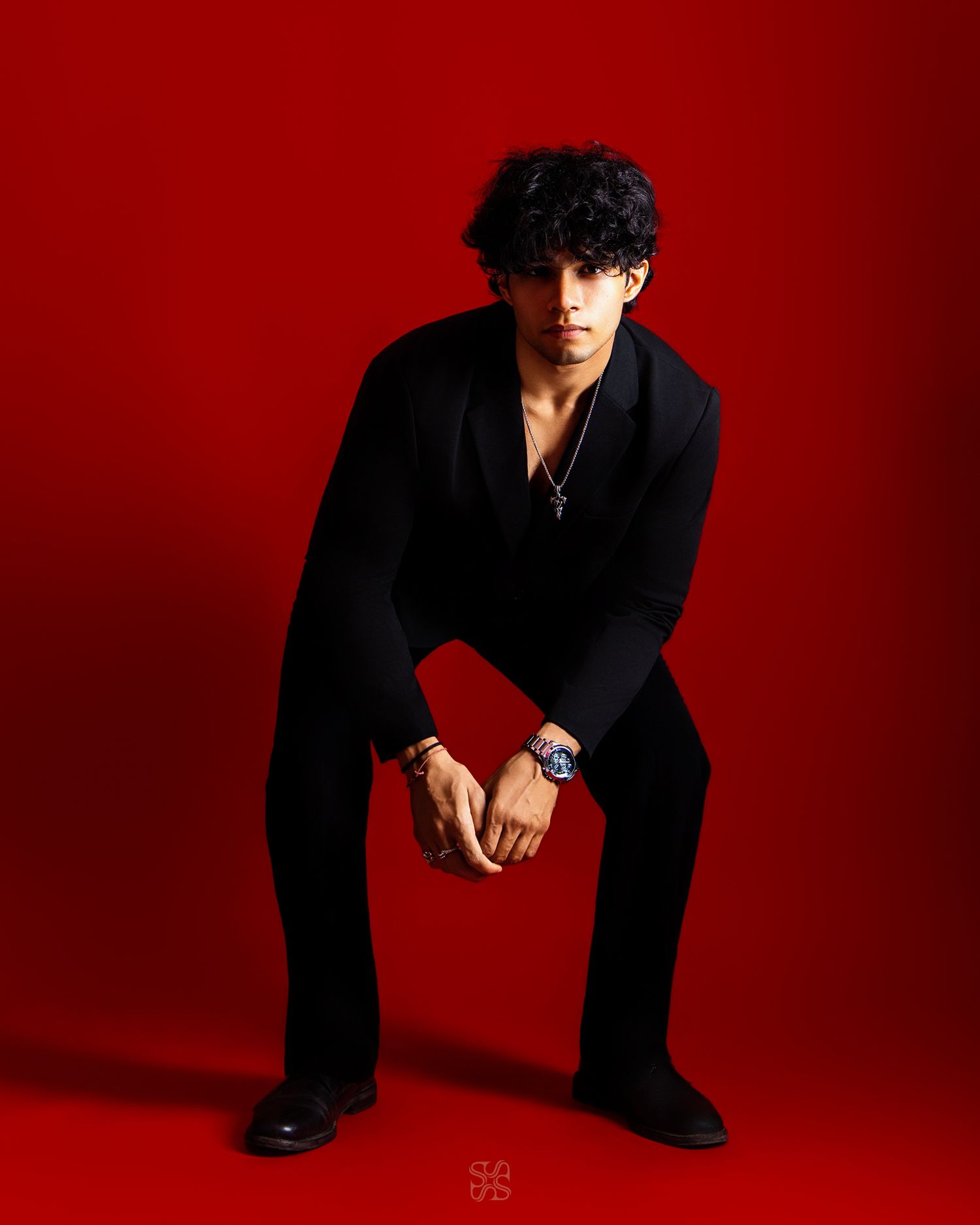 Young man in black outfit poses confidently against a bold red background, wearing a blue watch and necklace.
