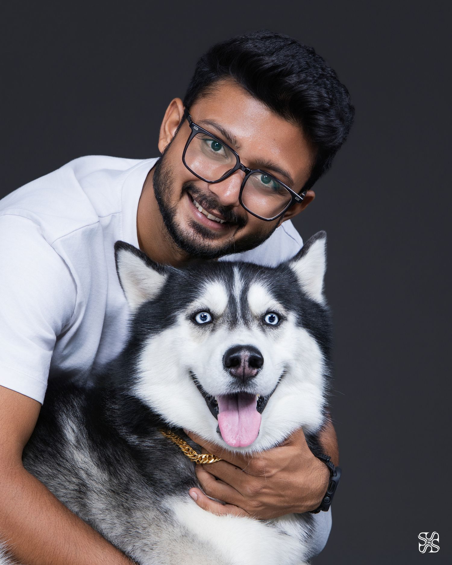 A smiling man with glasses hugging a blue-eyed Siberian Husky dog against a dark gray background.