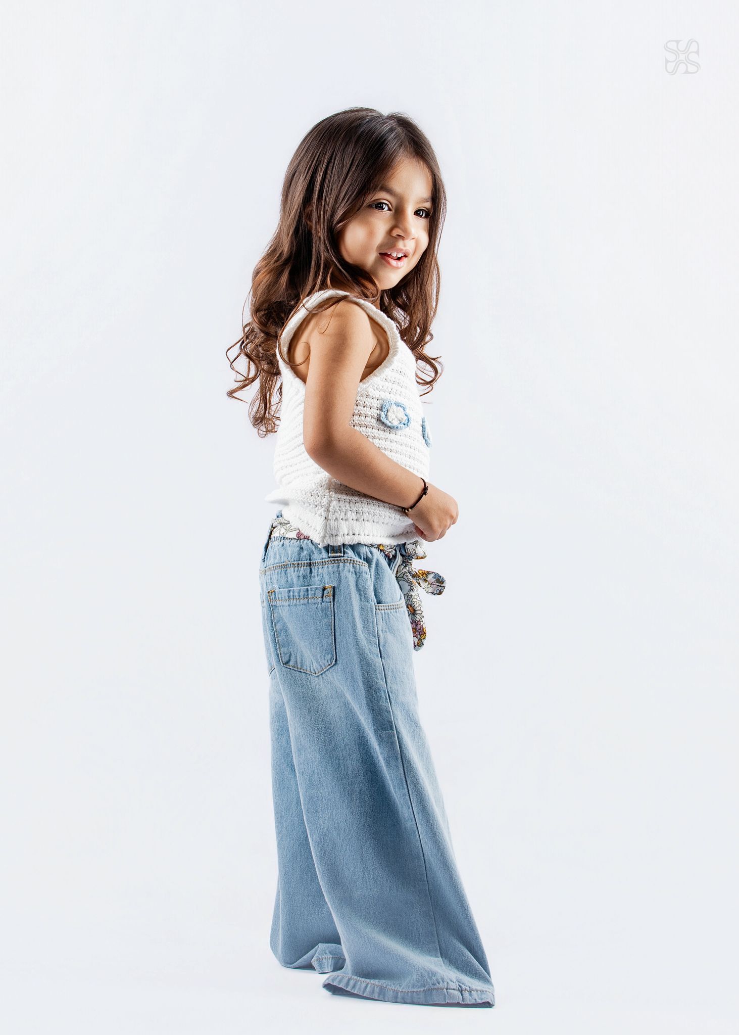 Young girl in white top and wide-leg light blue jeans posing sideways, smiling over shoulder on white background.