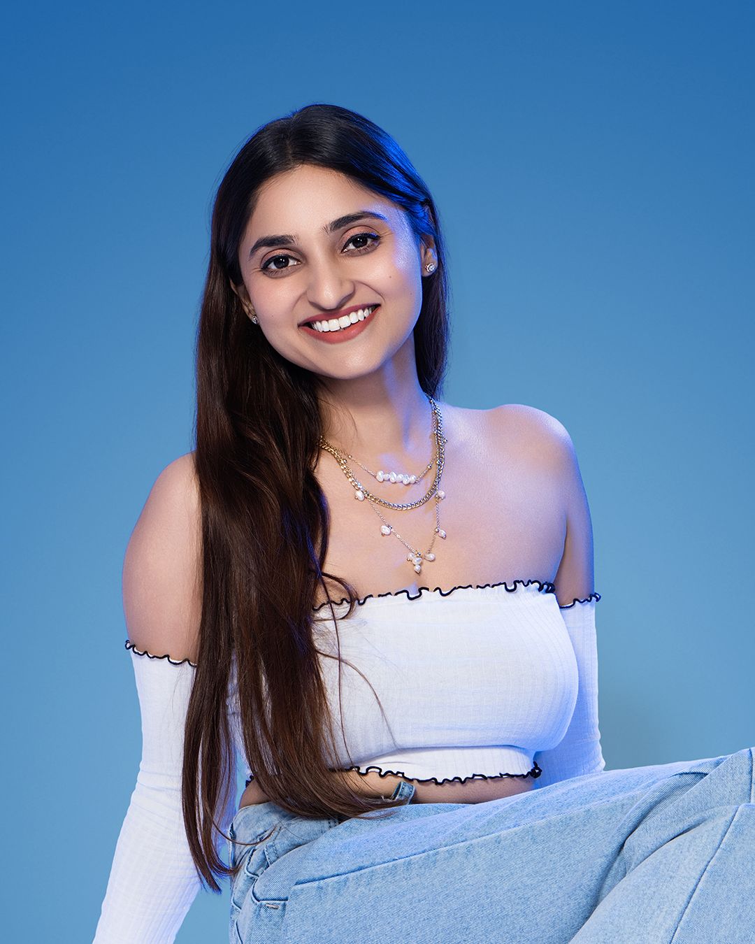 Young woman smiling in white off-shoulder top and layered necklaces against a blue background.