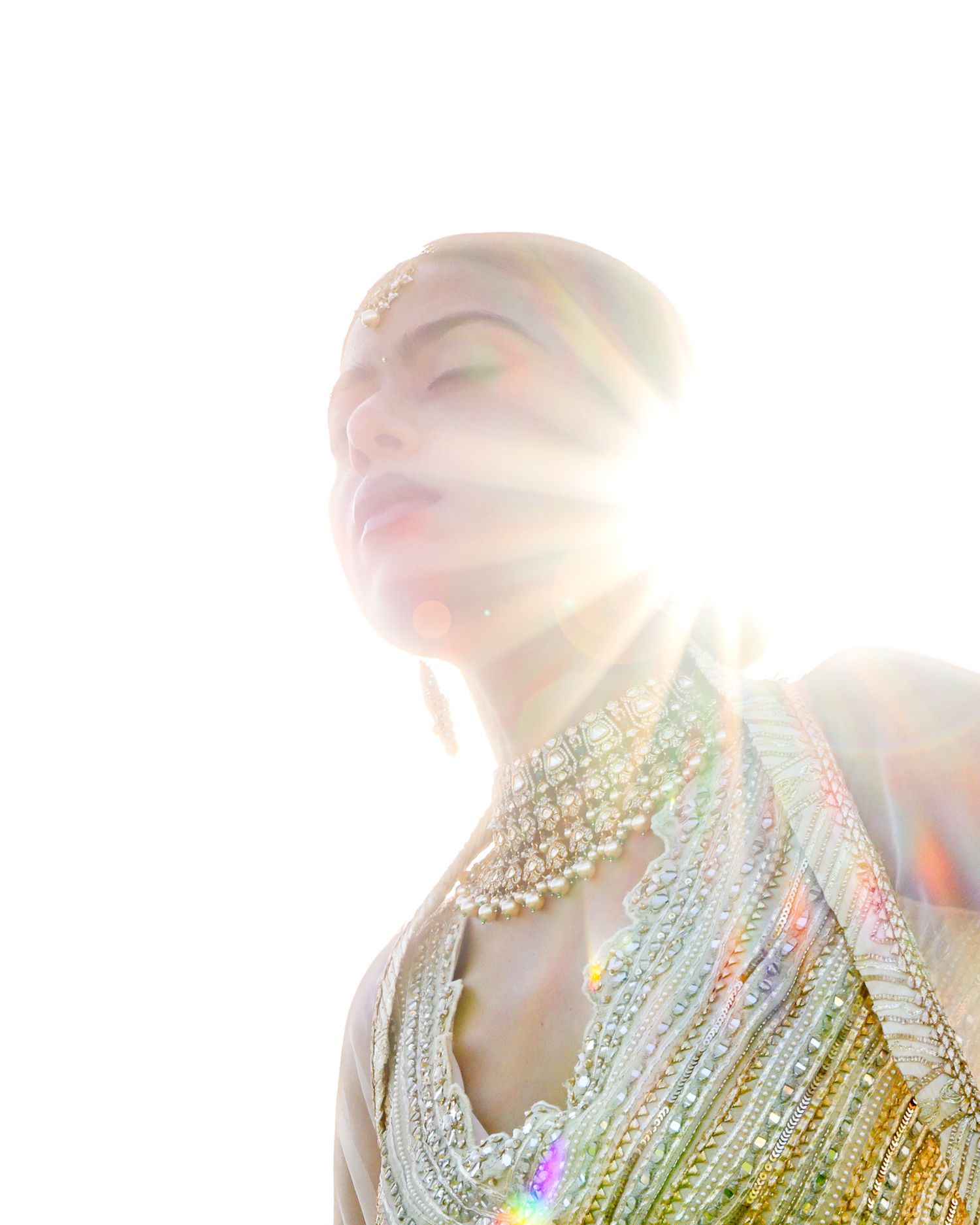 A woman in ornate beaded bridal jewelry poses in radiant sunlight with colorful lens flares illuminating her elegant attire.