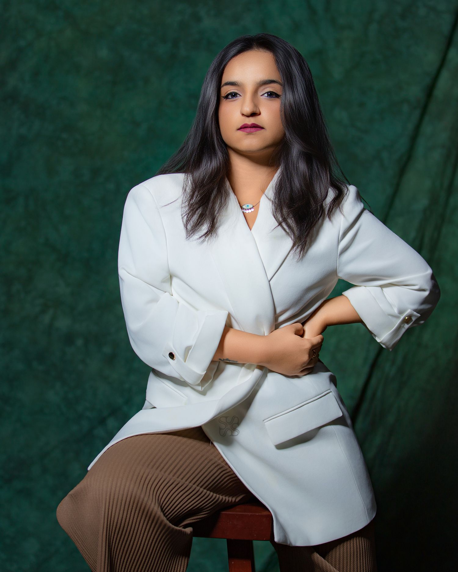 A young woman in a white blazer and brown striped pants sits confidently on a stool against a green backdrop.