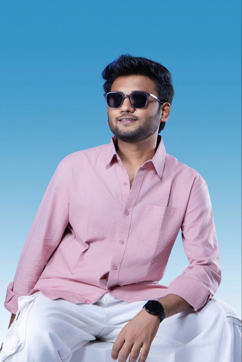 A person in a pink button-down shirt and white pants poses outdoors against a bright blue sky background.