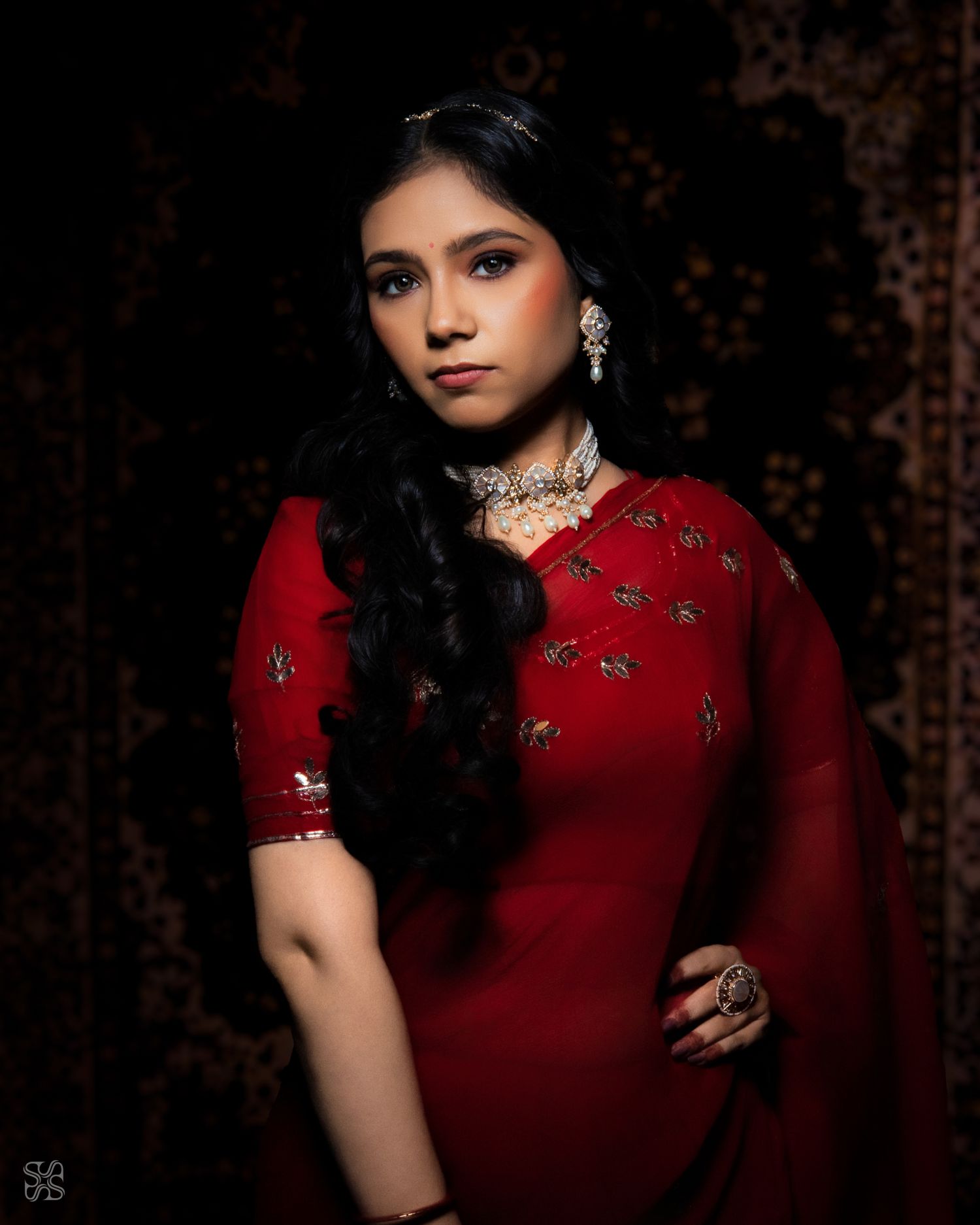 Elegant woman in red embroidered saree with silver choker and ring posing against a dark dramatic background.
