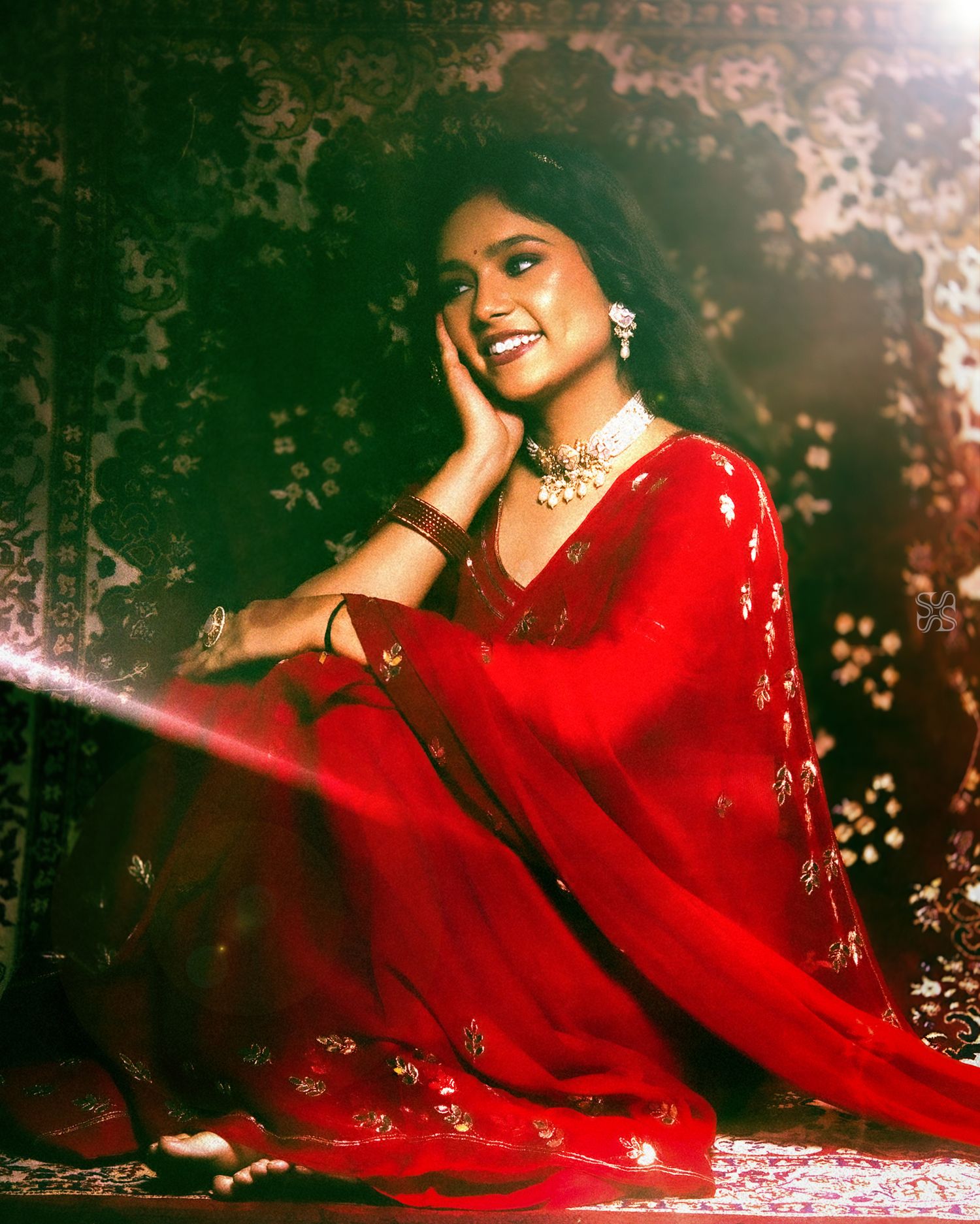 A smiling woman in a red embroidered saree and gold jewelry poses gracefully on the floor in dramatic lighting.