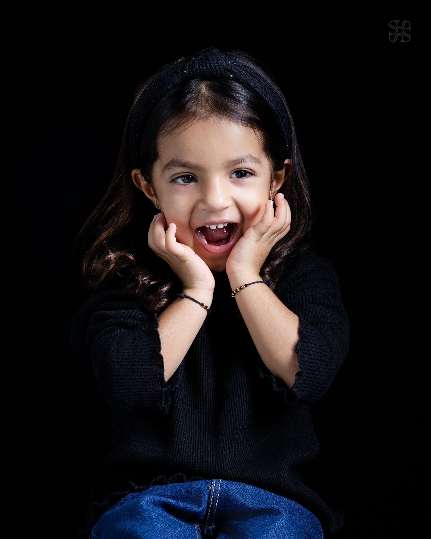 A joyful young girl in a black sweater poses with hands on cheeks, smiling broadly against a dark background.