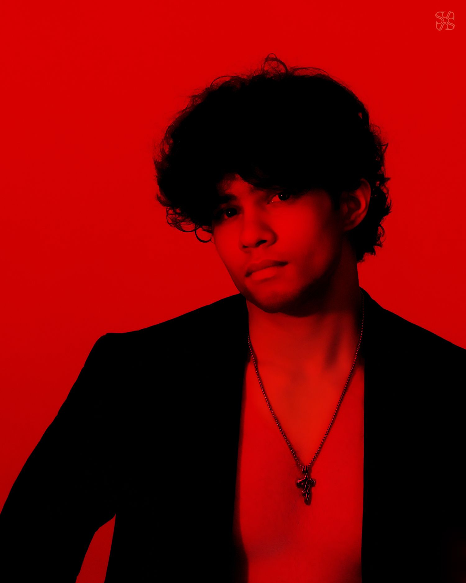 Young person with curly dark hair wearing a black blazer and cross necklace posed against a bold red background.