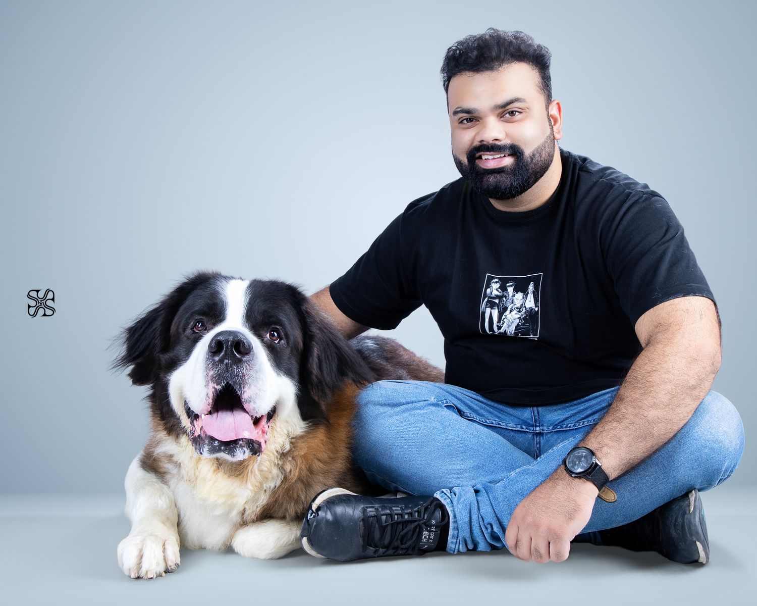 Man in black t-shirt sitting cross-legged beside a large Saint Bernard dog against a gray studio background.