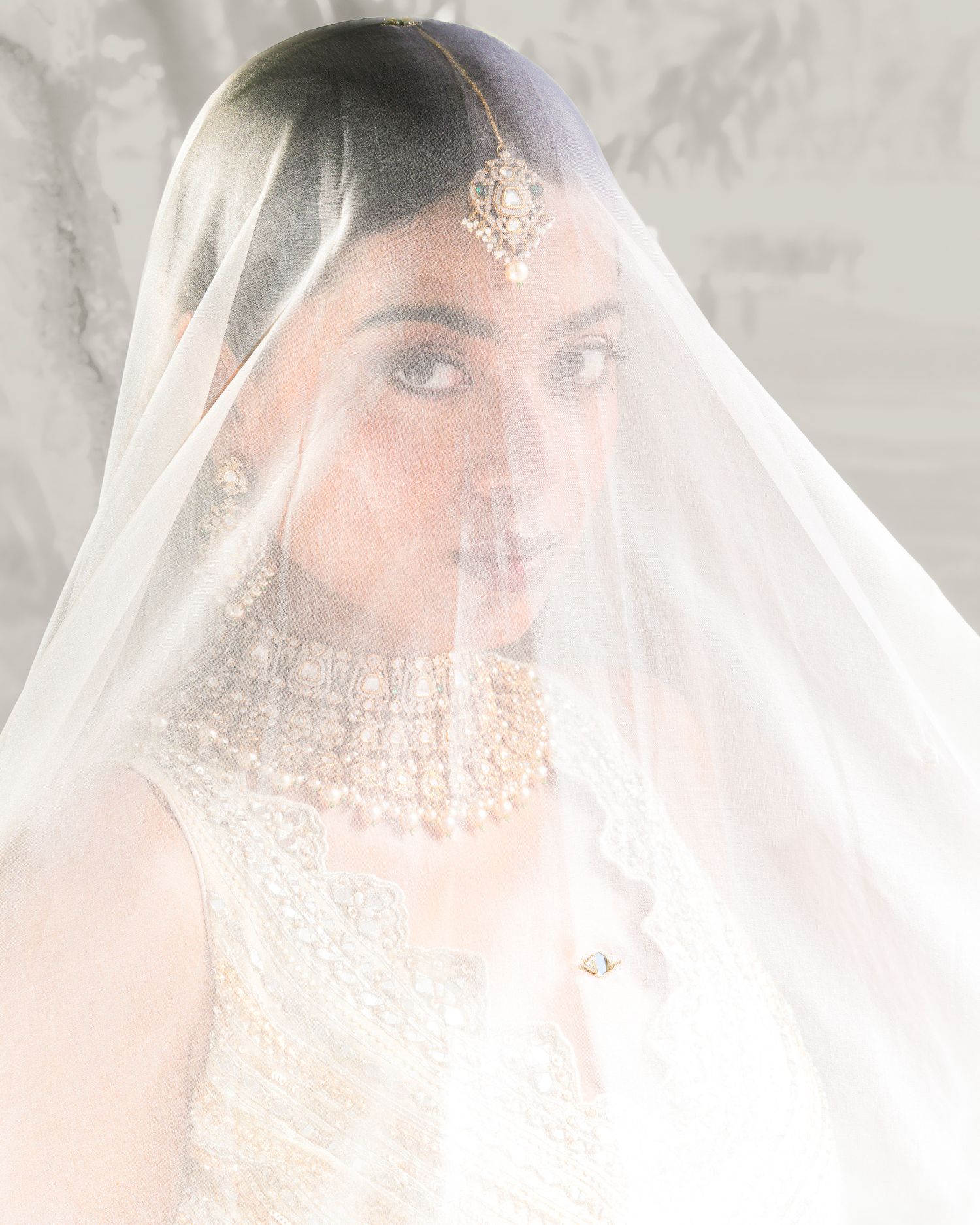 A bride draped in a white veil adorned with gold maang tikka and necklace.