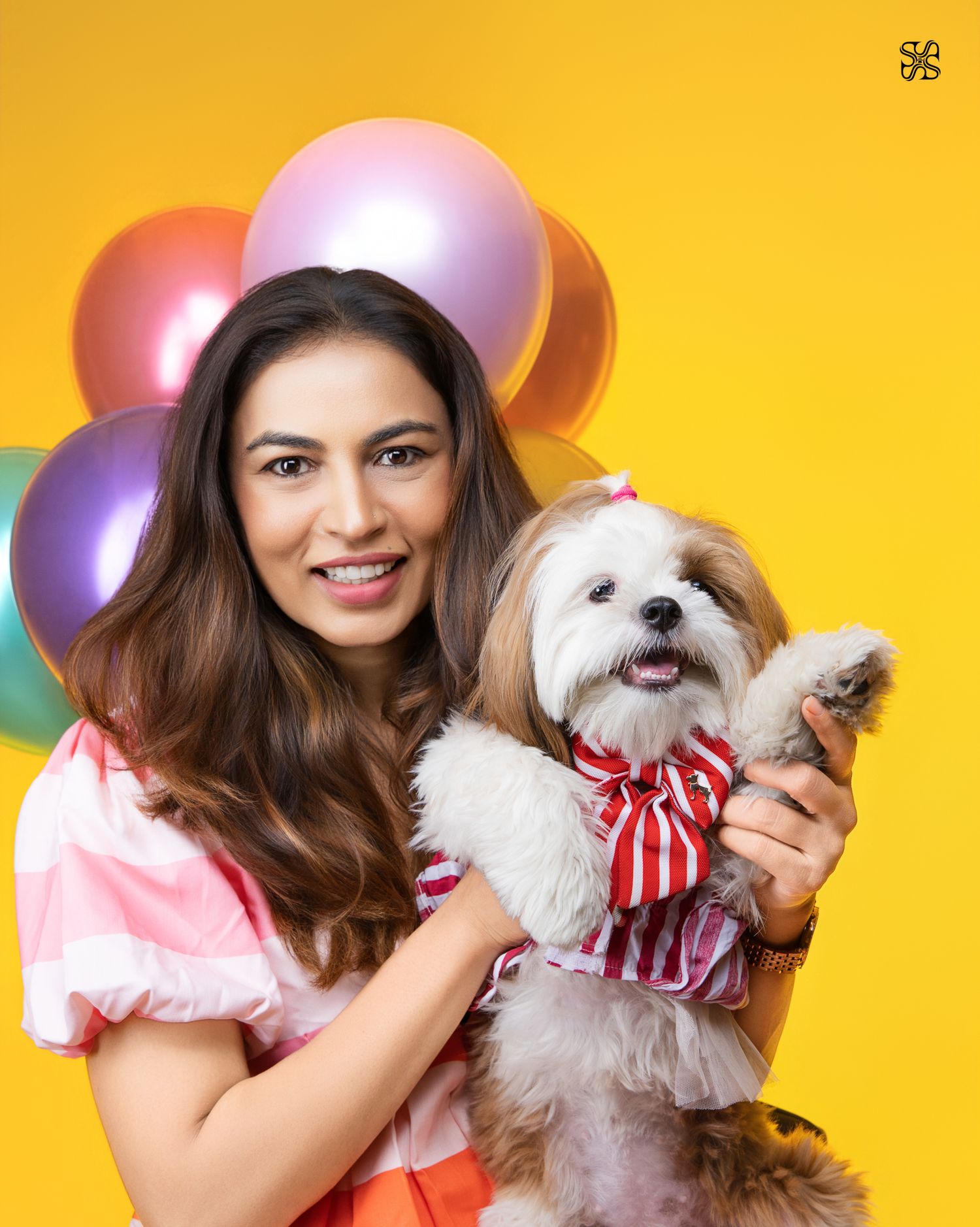 Woman smiling and holding a small dog wearing a red bow tie, with colorful balloons on a yellow background.