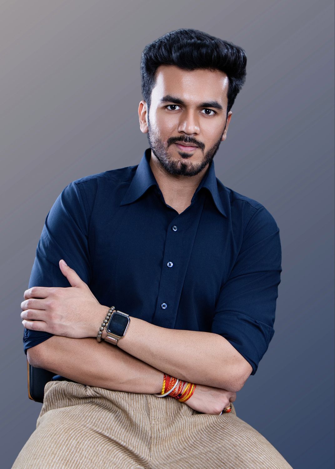 Young man in navy shirt with crossed arms, wearing smartwatch and bracelets, posing confidently against gray background.