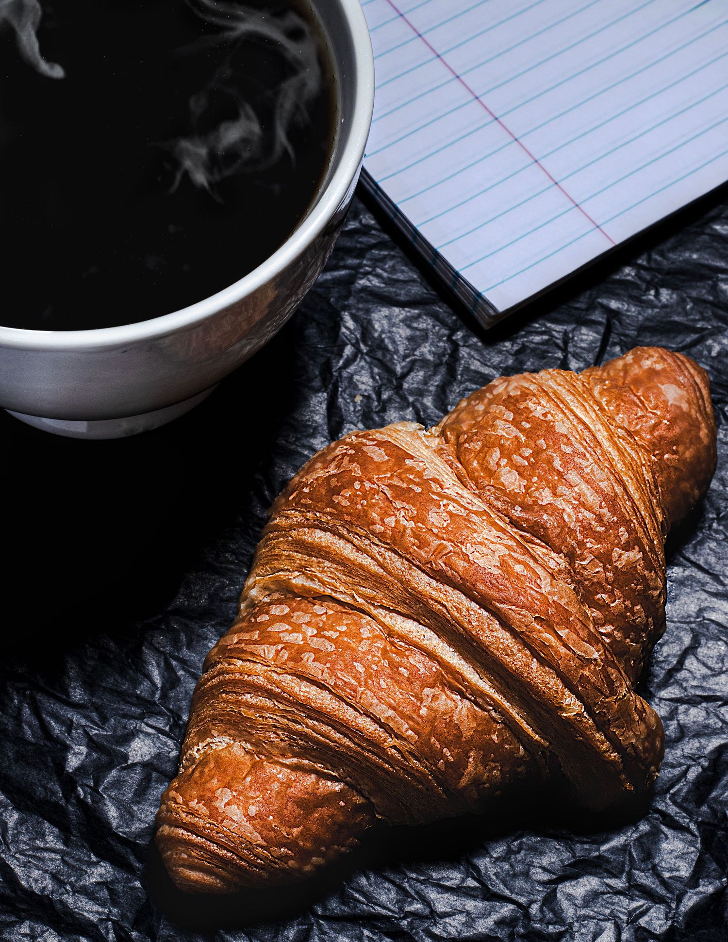 A freshly baked golden brown croissant photographed against a dark background with coffee cup and notepad visible.