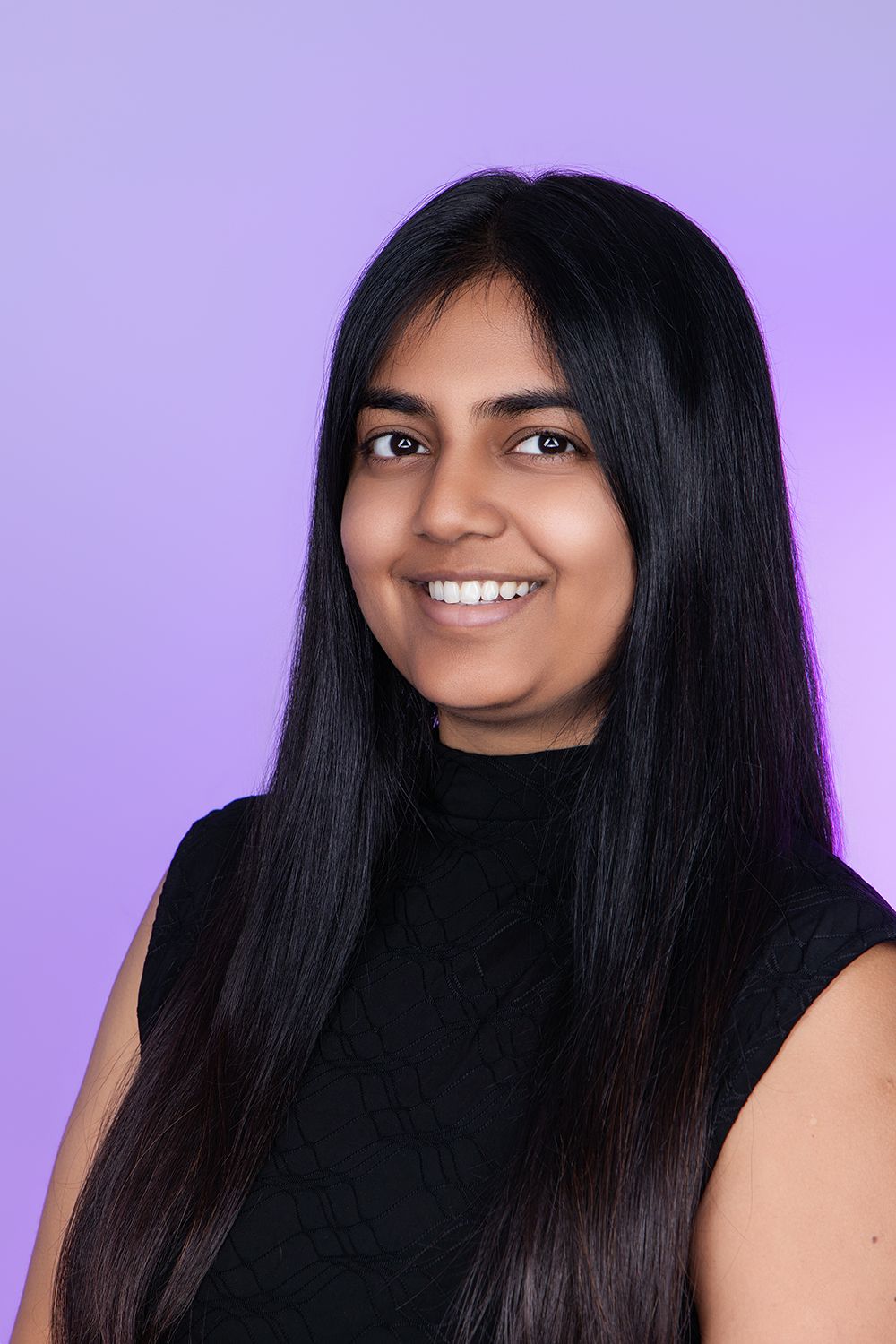 Smiling young woman with long black hair wearing a black sleeveless top against a purple background.