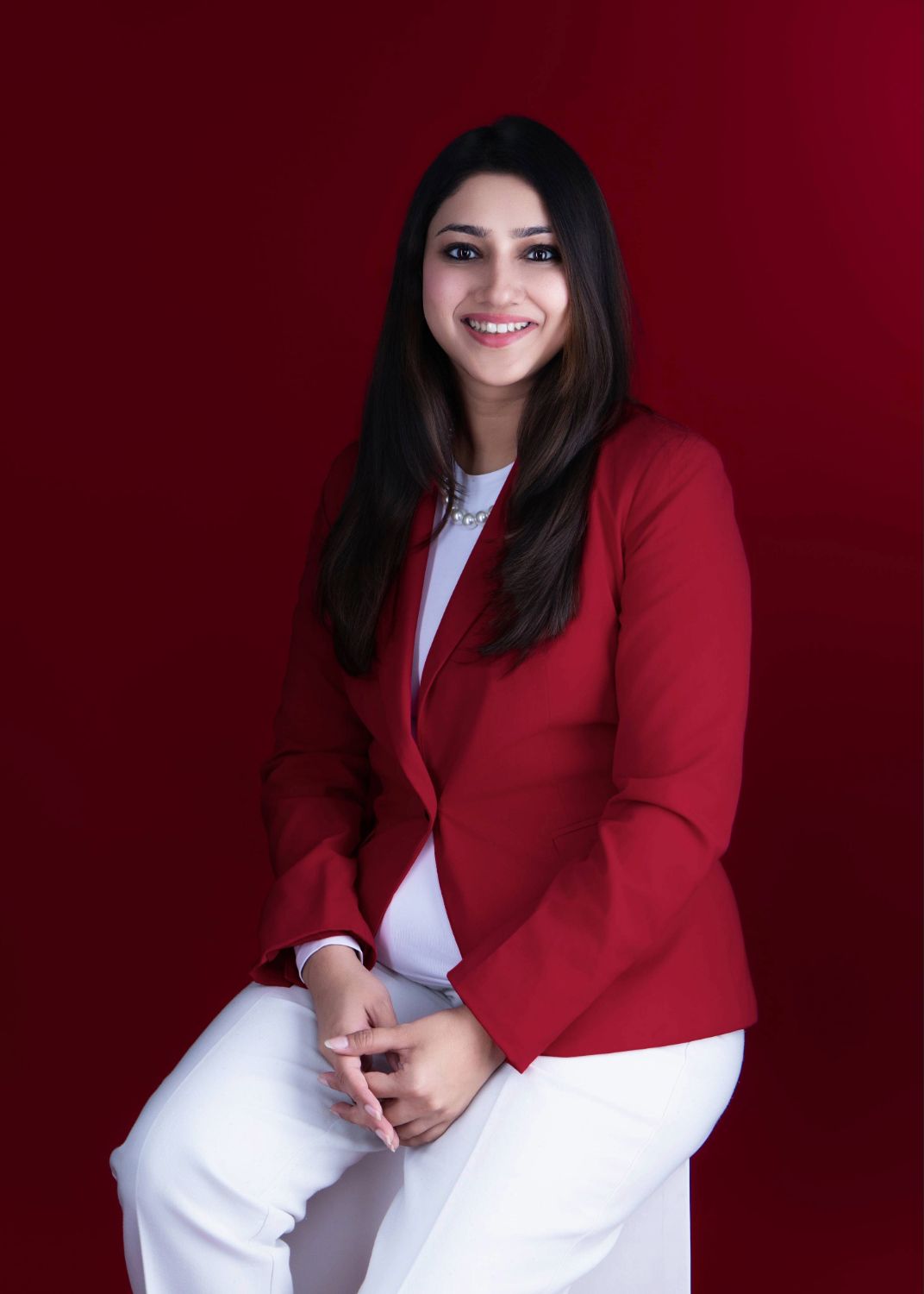Professional portrait against a red background showing a person in a red blazer and white outfit sitting with clasped hands.
