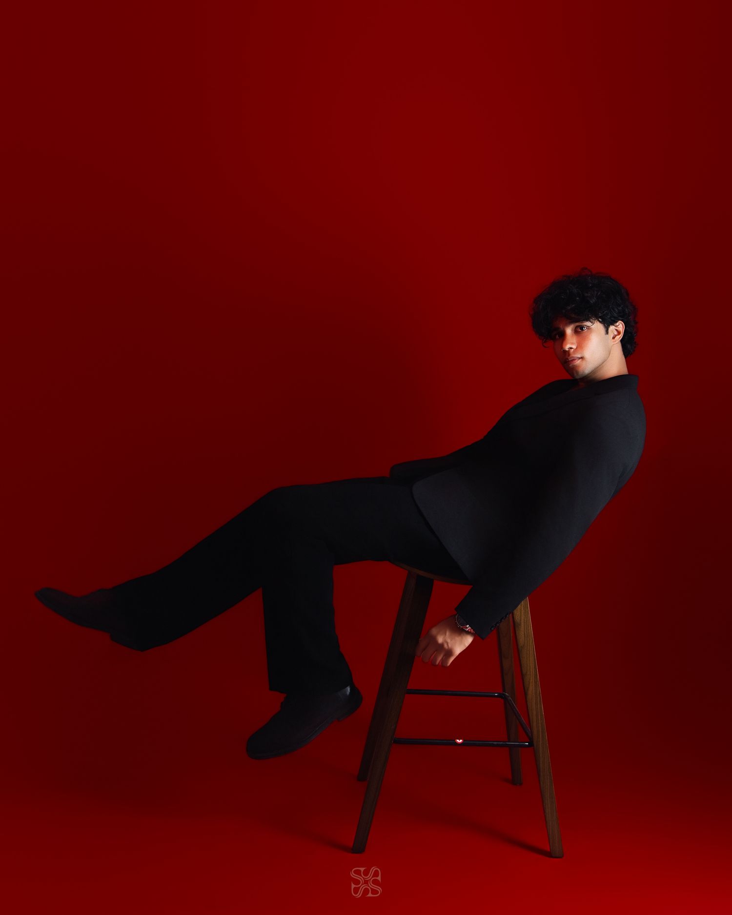 Young man in all-black outfit posed on wooden stool against bold red background, looking over shoulder.