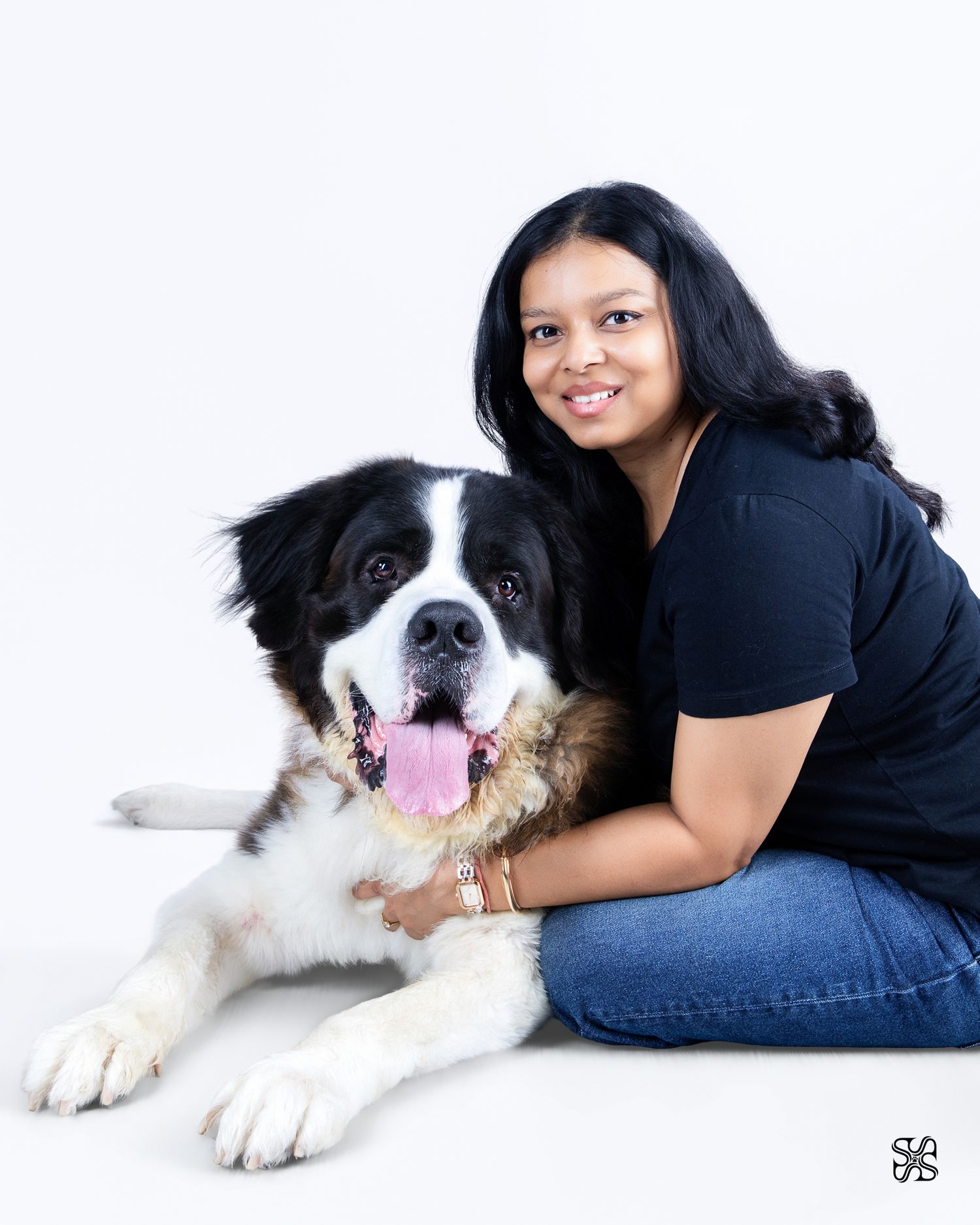 A smiling woman in a black shirt hugs a large black and white Saint Bernard dog against a white background.