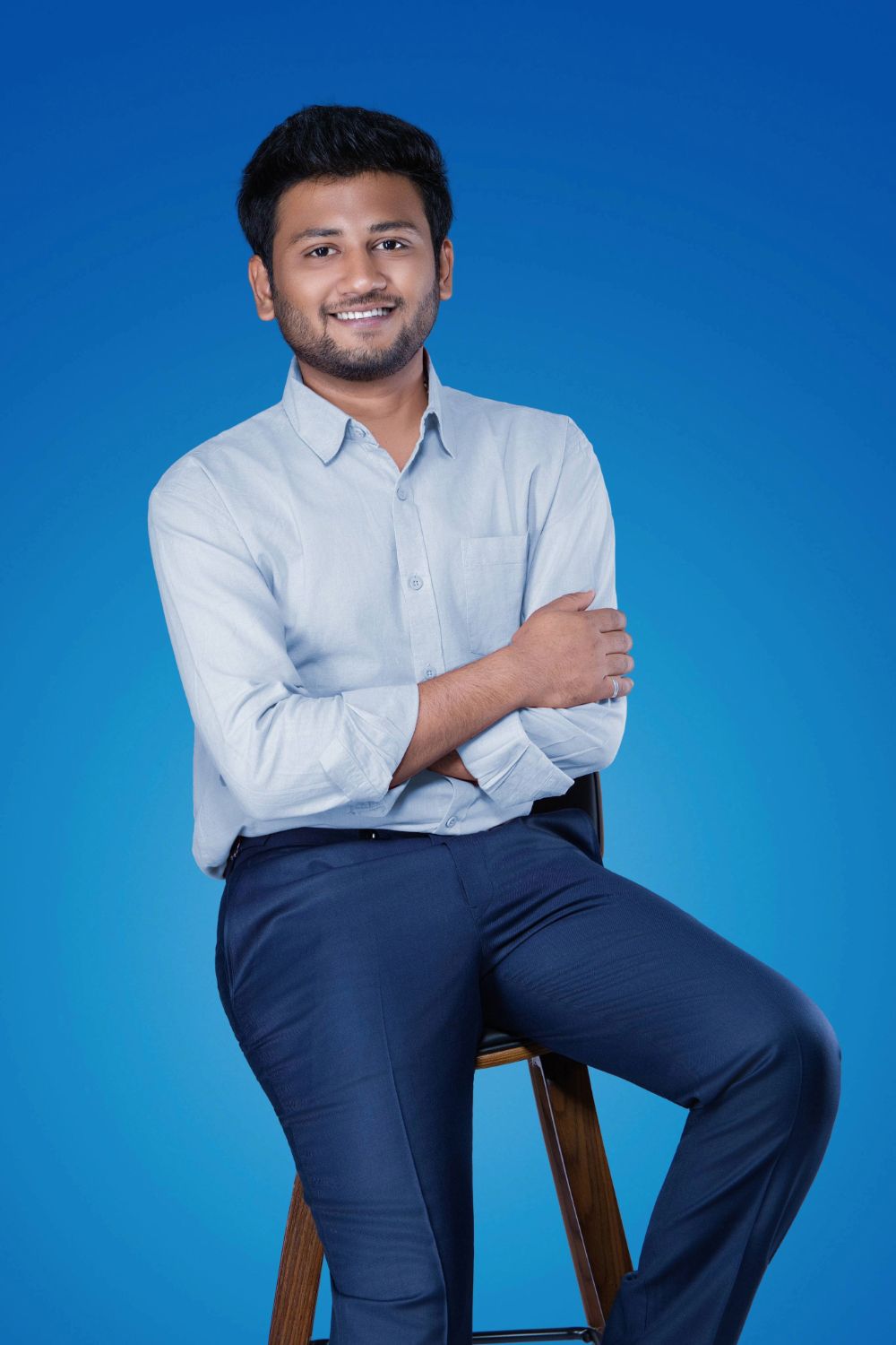 Young Indian man in light blue shirt and navy pants sitting on stool, smiling confidently against blue background.