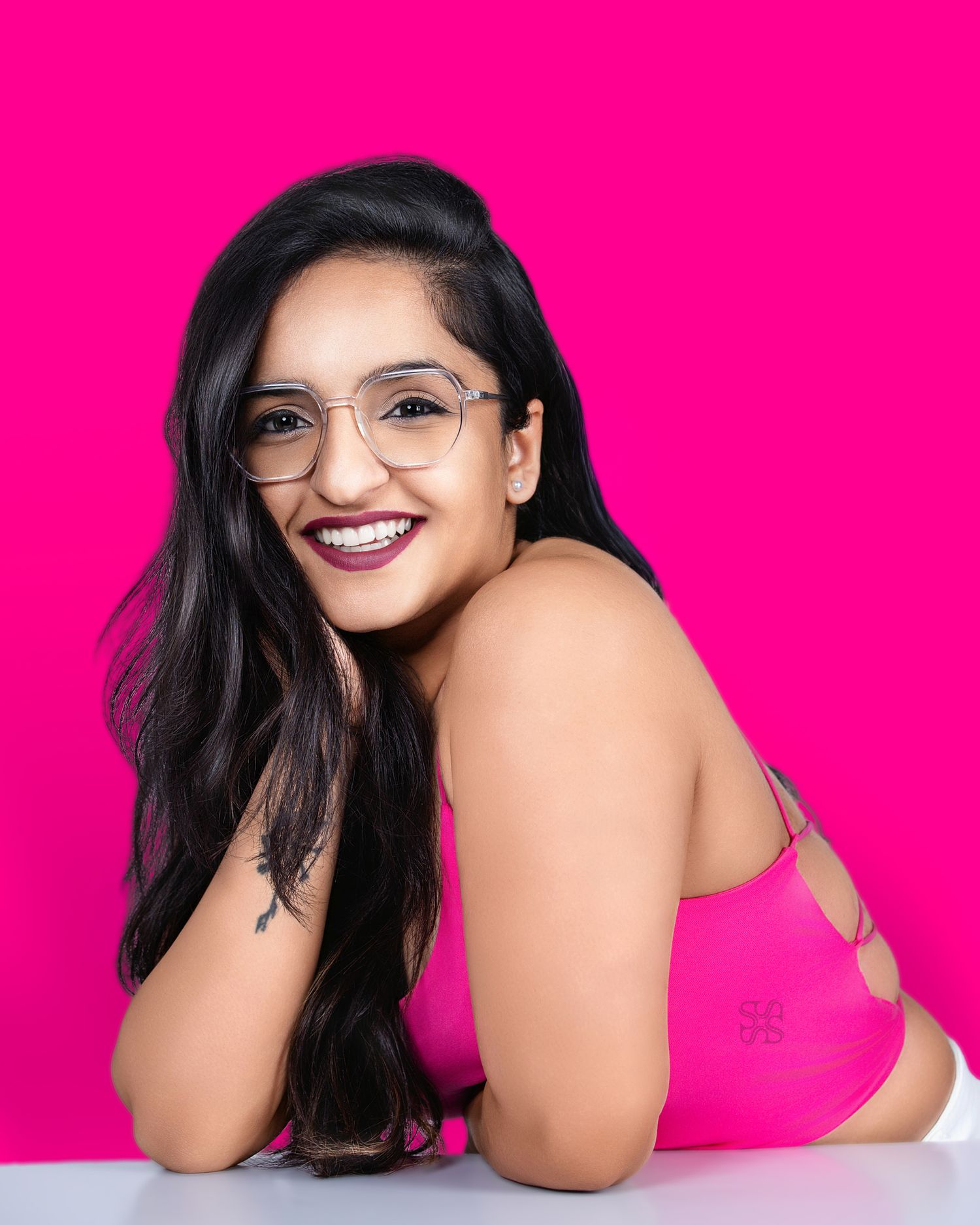 Smiling young woman in pink outfit and glasses posing against a vibrant pink background.