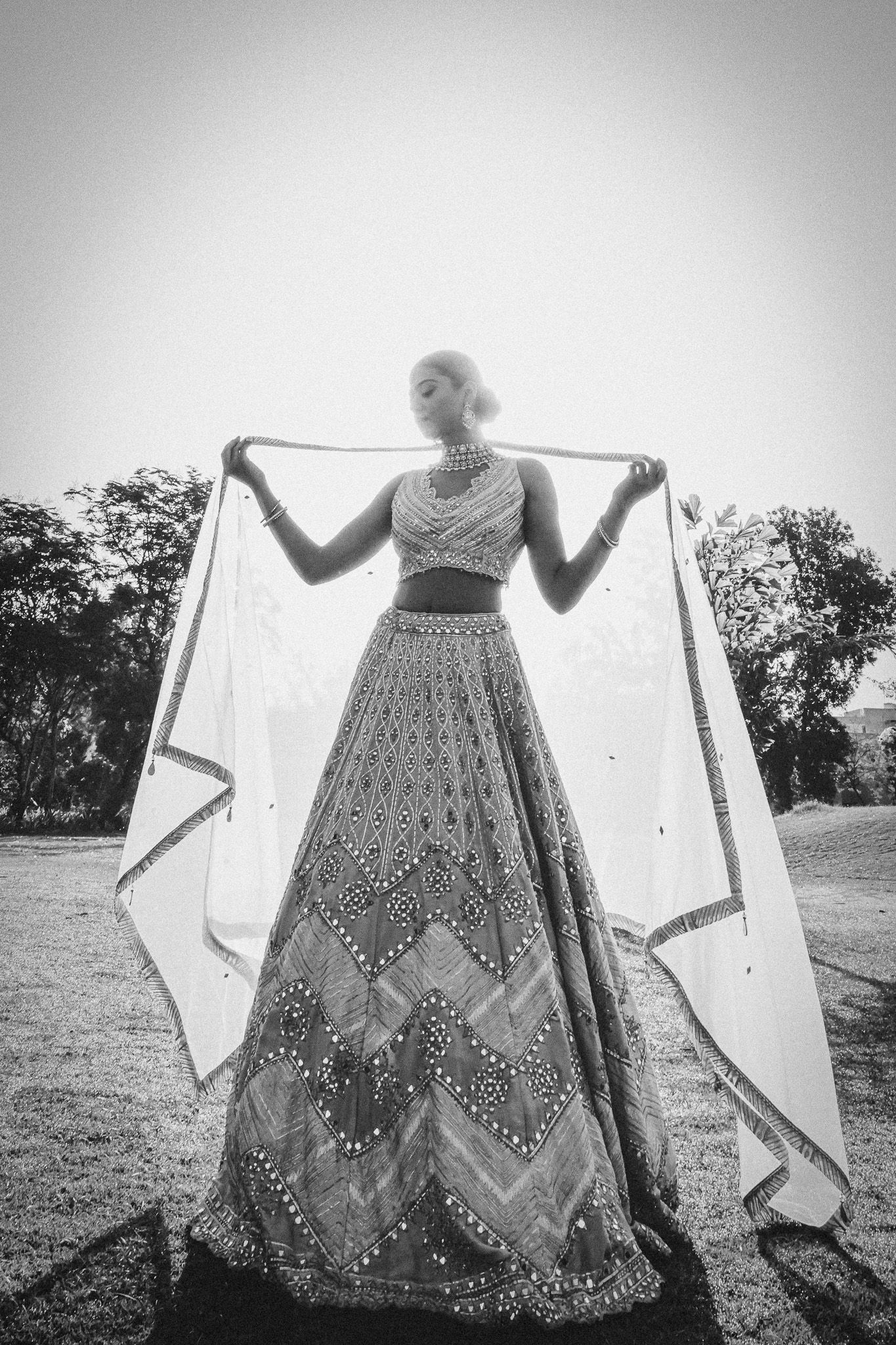 Black and white photo of a woman in an embroidered lehenga holding a dupatta wide open outdoors.
