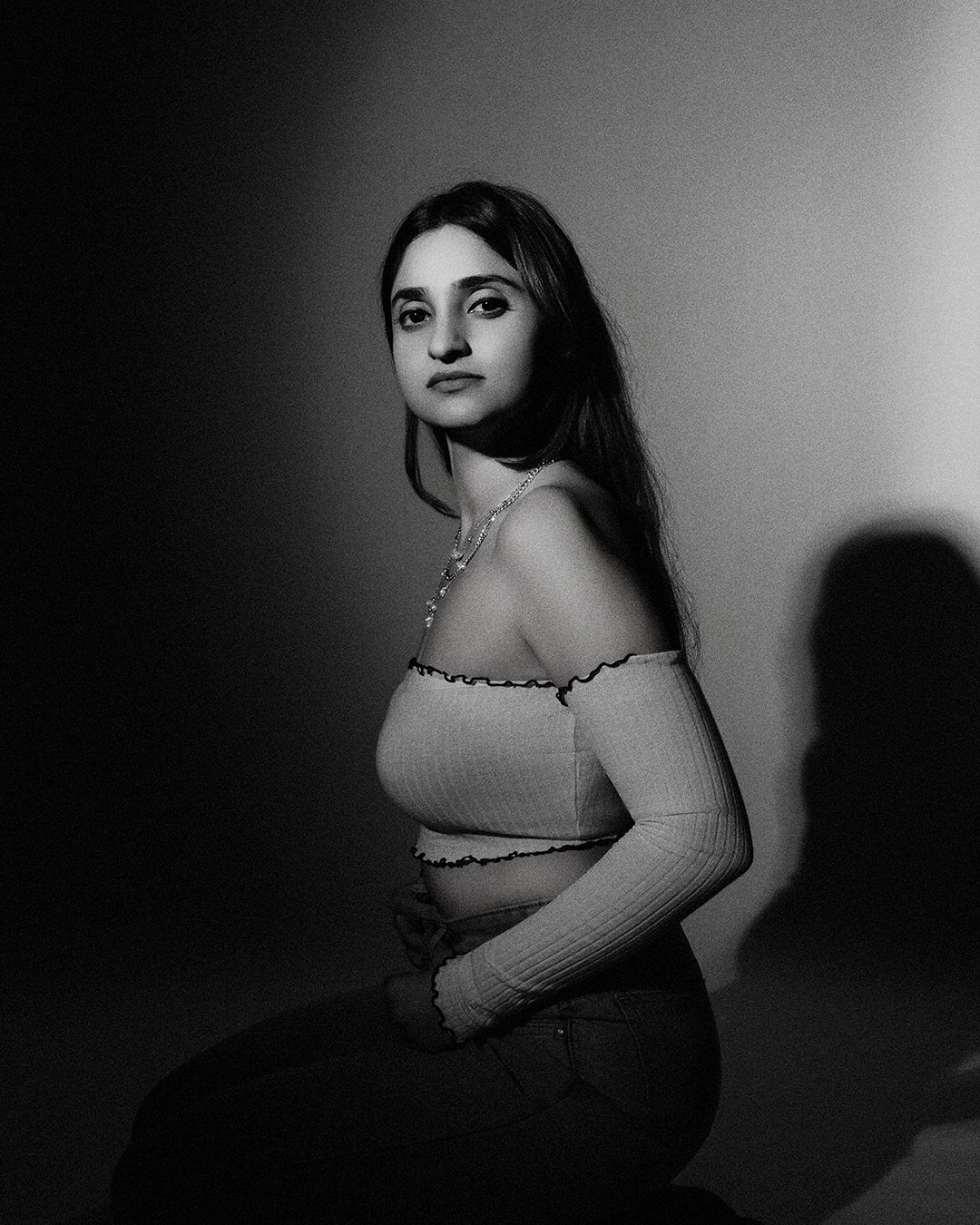 Black and white portrait of a young woman in an off-shoulder crop top, posing against a softly lit wall.