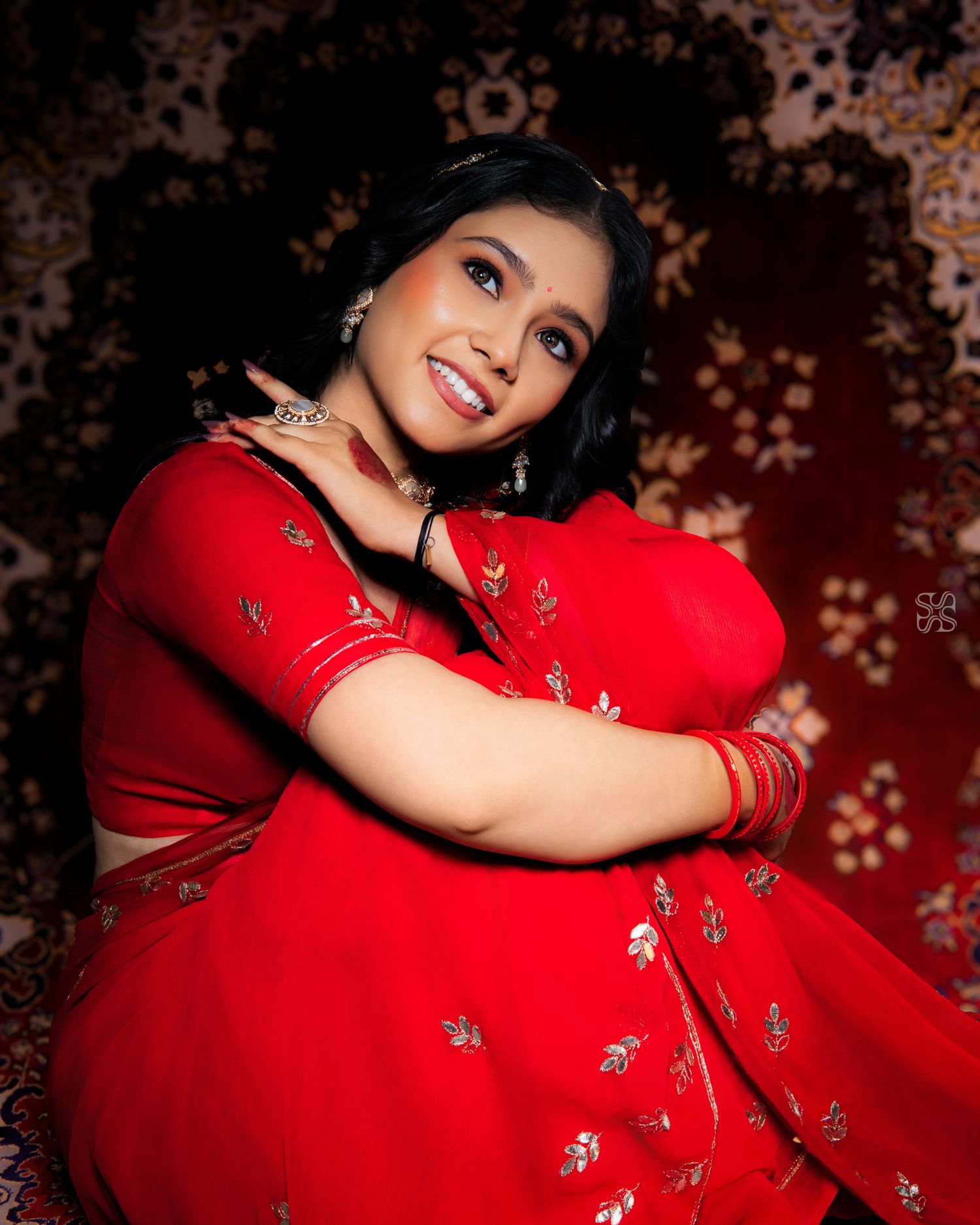 Smiling woman in red embroidered saree with silver jewelry posing gracefully against a dark floral background.