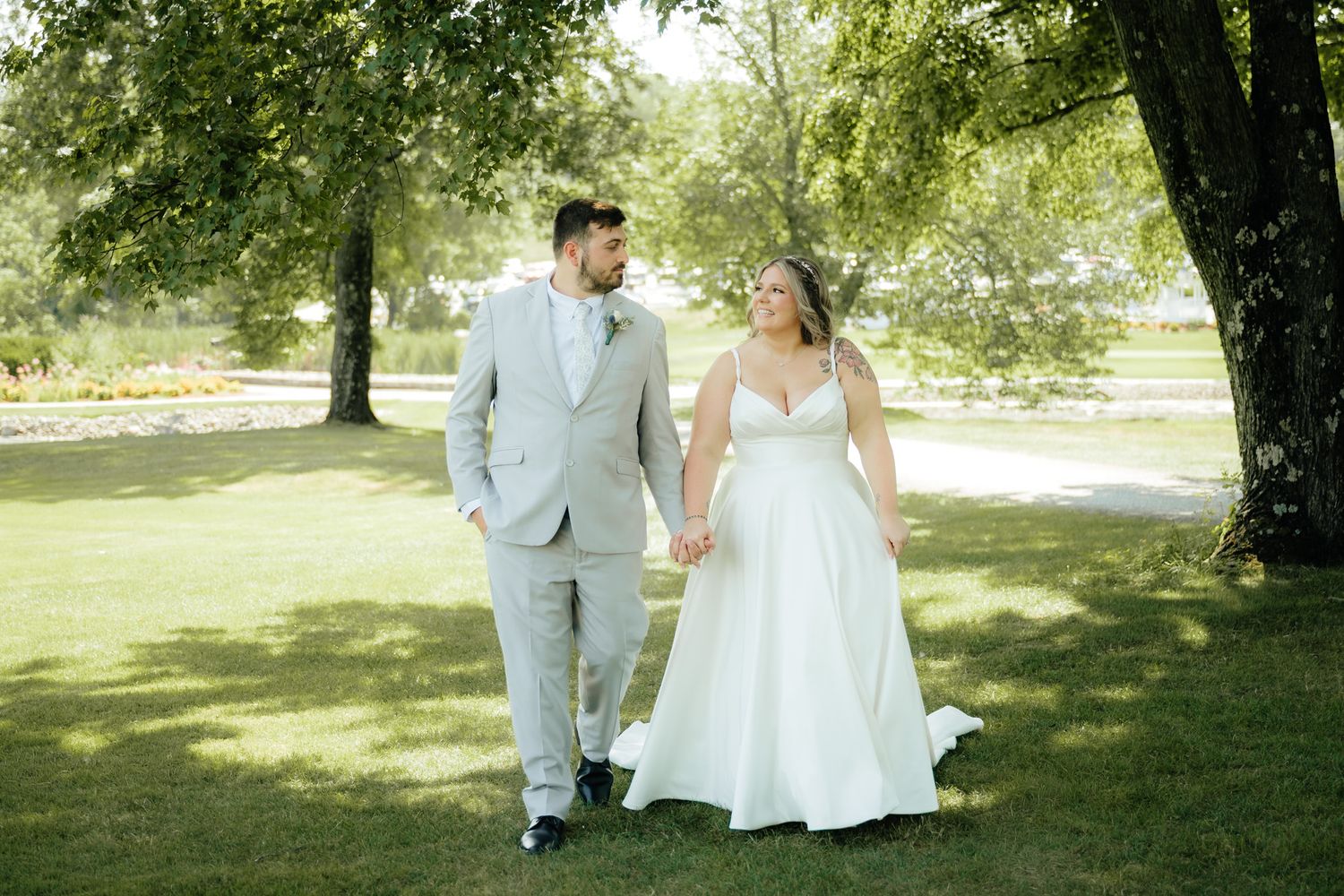 Bride and groom walking hand in hand through lush green park on sunny summer wedding day.